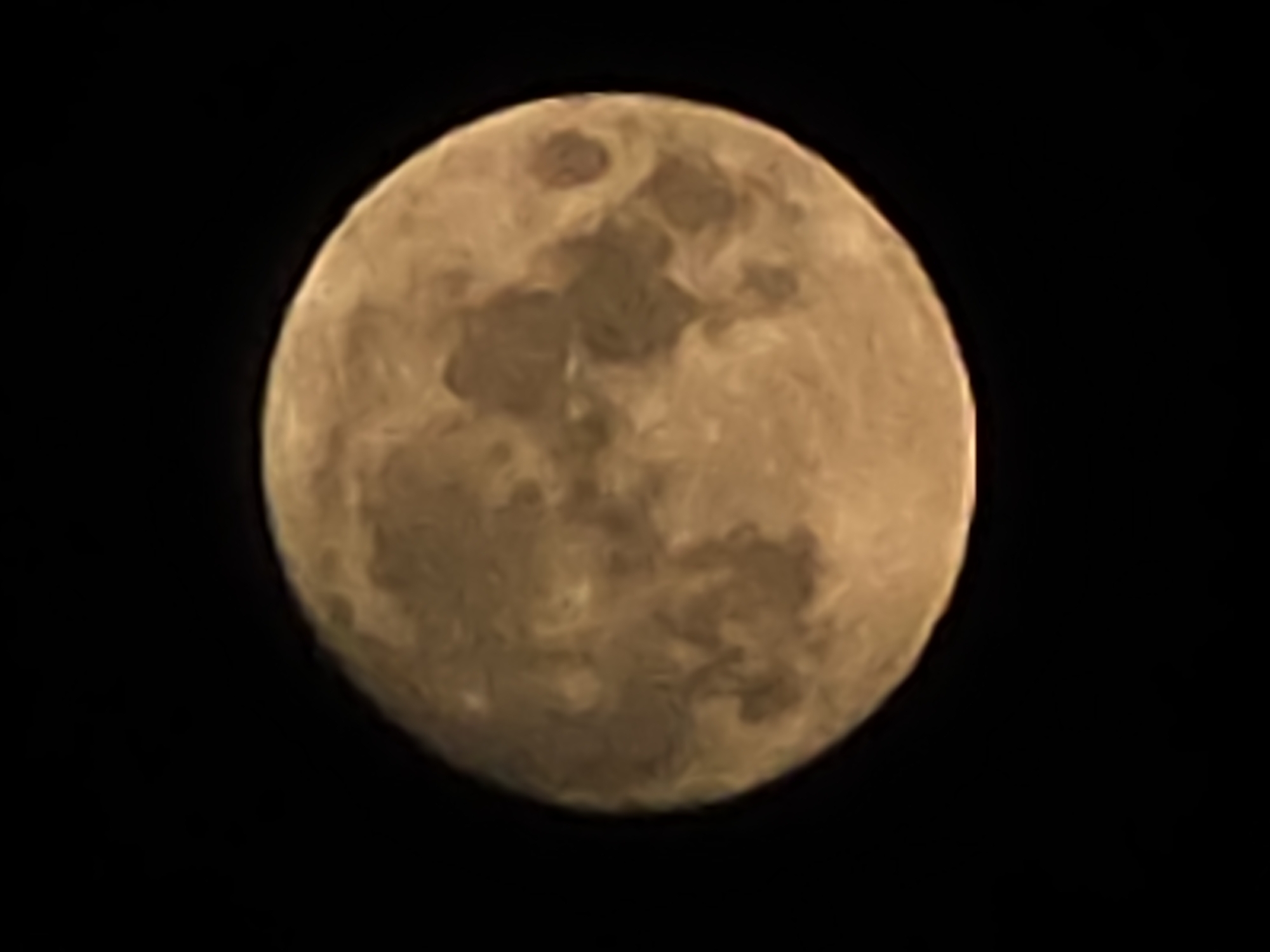 A close-up image shows the full moon against a black sky. The moon has a tan hue and visible surface features with darker and lighter patches creating a mottled appearance. The image has a slightly blurred quality.