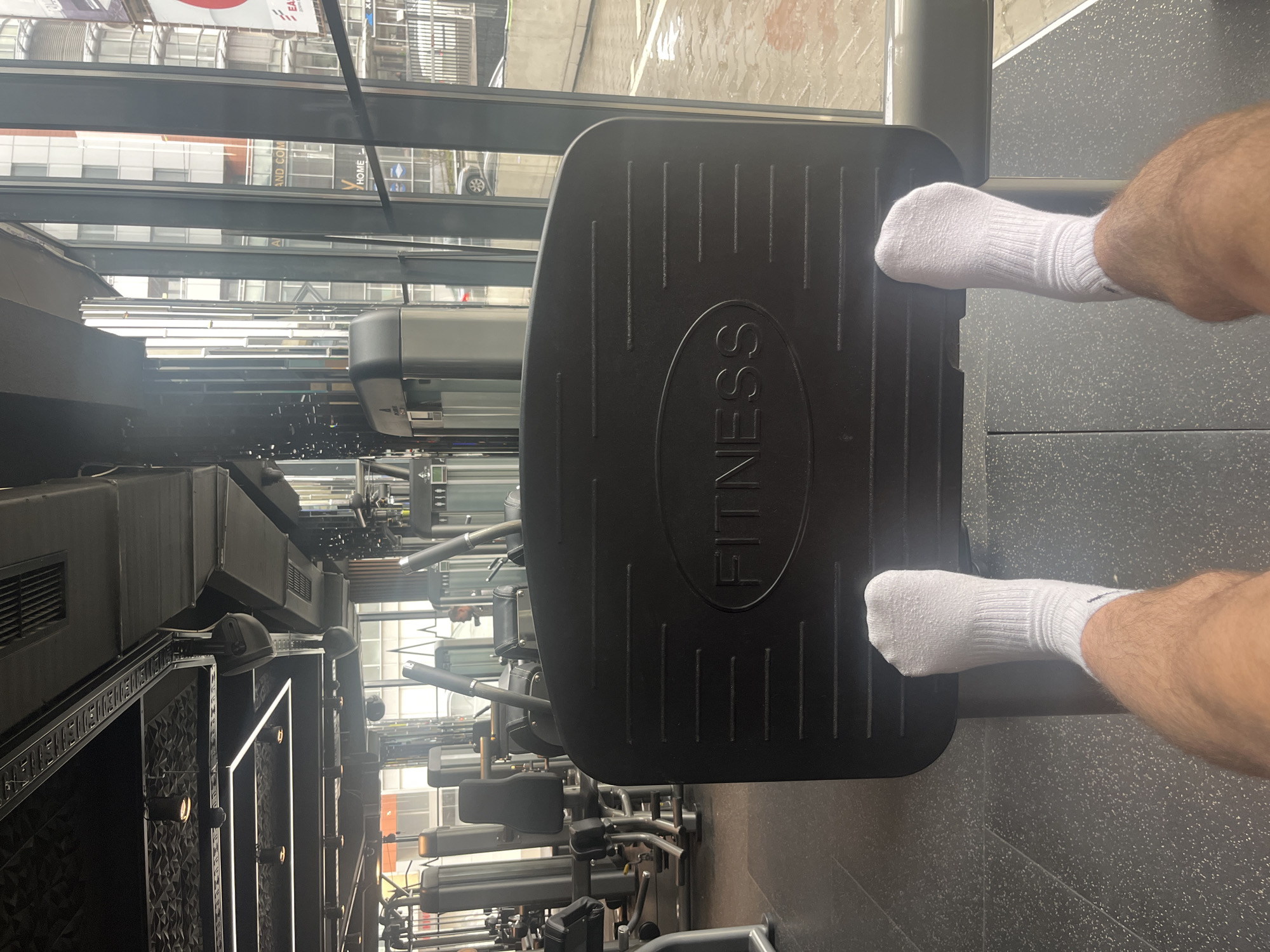 The image depicts an indoor gym setting. The camera focuses on a leg press machine, with a person's lower legs and feet, clad in white socks, resting on the footplate. The footplate is black and has the word "FITNESS" vertically inscribed on it. The gym equipment, including rows of weight machines, extends into the depth of field. Large windows reveal an urban environment. The location is the city of Sofia, Bulgaria.