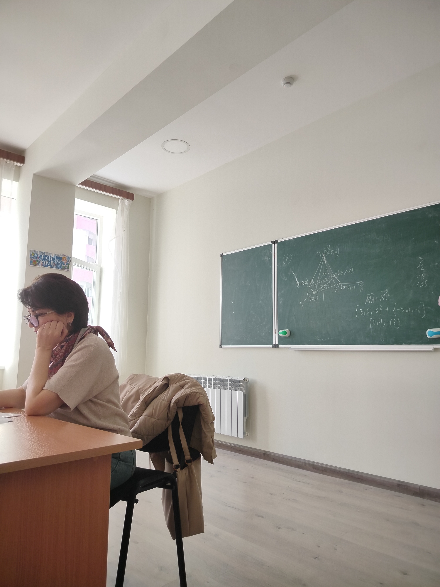 The image shows a woman sitting at a desk in a classroom setting. She has short dark hair, wears glasses, and is wearing a light-colored top with a patterned scarf. Her hand is resting on her face as if she is thinking. In front of her is a wooden desk. A jacket is draped over the back of the chair.
Behind the woman, there is a window, revealing what appears to be a multi-story building, suggesting an urban environment, which may be in Yerevan, Armenia. To the right is a blackboard covered in math problems and geometric diagrams. A white radiator is on the wall beneath the blackboard. The room has light-colored walls and a light-colored floor. The classroom appears to be clean and well-lit.