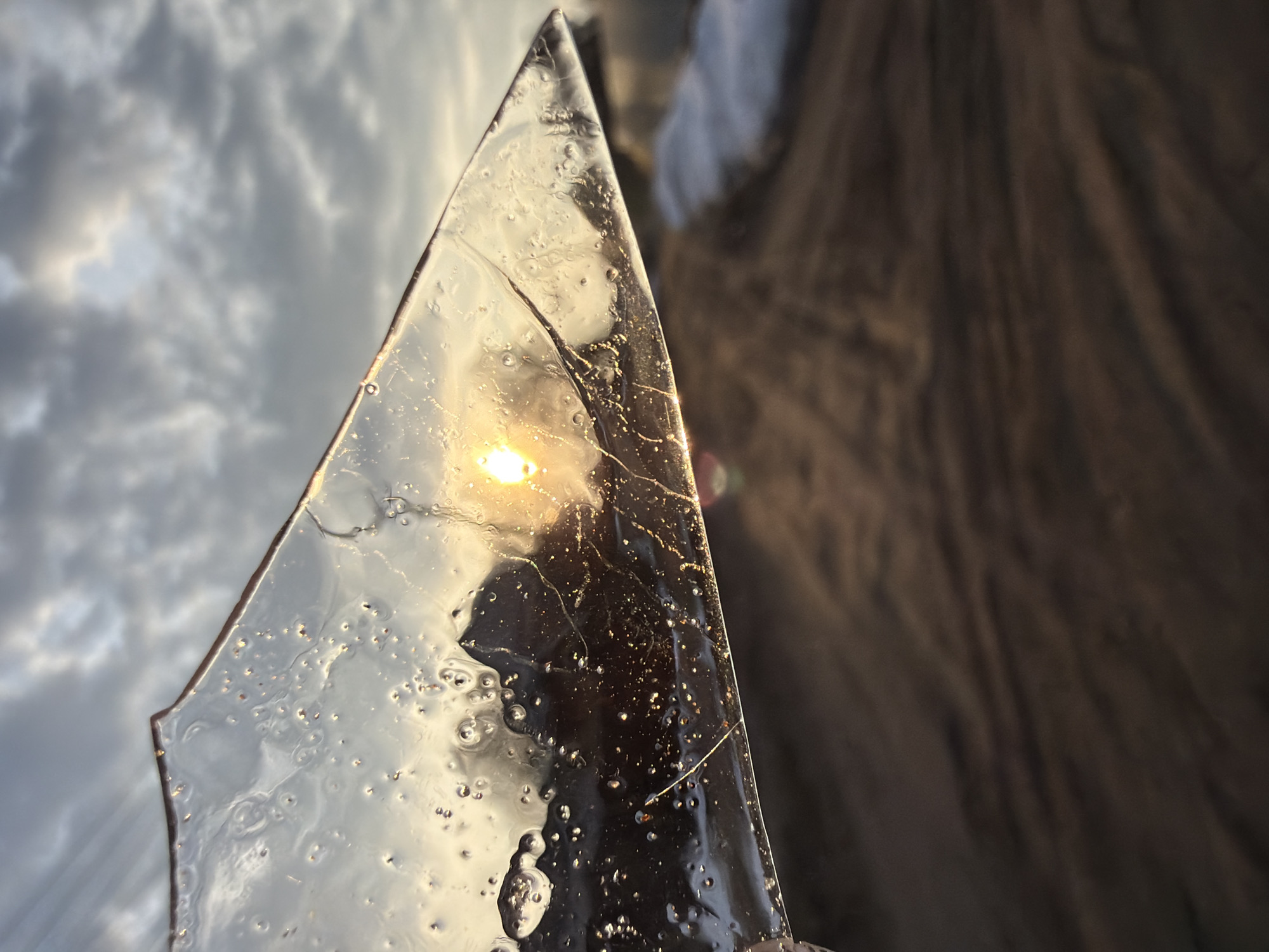 The image presents a close-up view of a roughly triangular piece of transparent material, possibly glass or clear plastic, held against the sky. The material contains numerous air bubbles and appears slightly cracked in places. Light, likely from the sun, shines through the material, creating a bright reflection within it. The sky is visible in the background, showing a mix of light and dark clouds. To the right of the transparent material is a blurred background that appears to be a dark, textured surface. No people, specific objects other than the material, actions, or interactions are visible. There is no way to verify the location as Chaikovsky, Russia from the image.