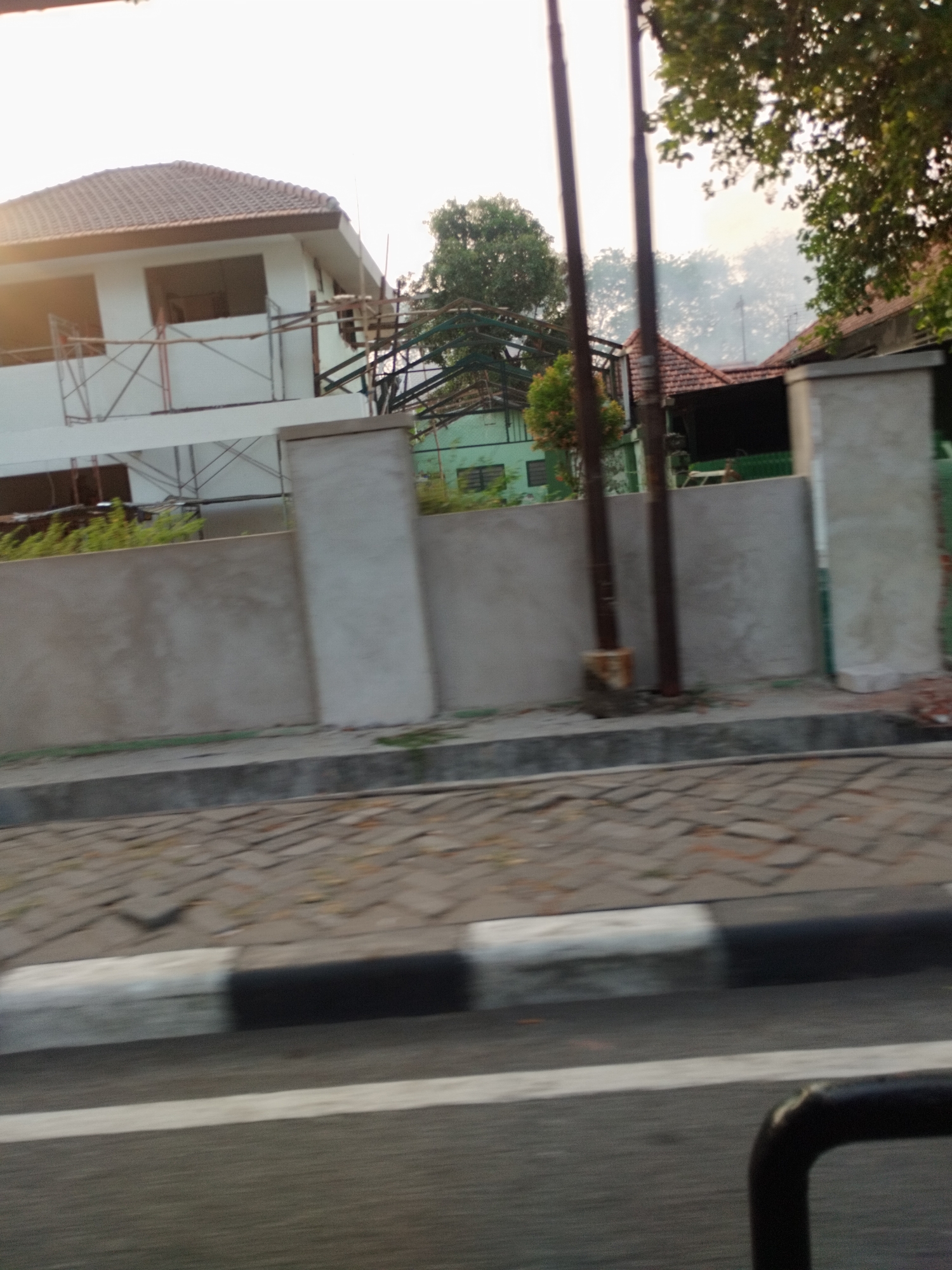 The image depicts a residential area in Surabaya, Indonesia, with a house under construction in the foreground. The house, painted white with a tiled roof, is partially covered with scaffolding, indicating ongoing building work. Adjacent to the house is a light green building with visible windows. A concrete wall runs along the front of the property, with two large concrete pillars as part of its structure. The street in front of the wall is paved with interlocking bricks, bordered by a curb with black and white markings. Trees and foliage are visible in the background, suggesting a semi-urban or suburban setting. The overall scene captures a moment in the development of a residential area in Surabaya.