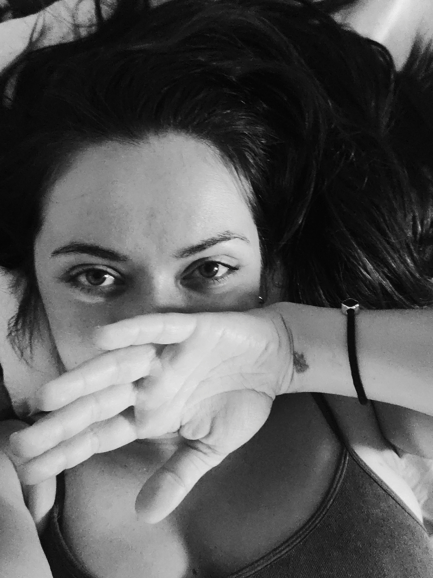 A black and white, close-up, high-angle portrait shows an individual, likely female, lying down and looking directly into the camera. The subject's dark hair is disheveled and fanned out around the head. Both eyes are open, dark, and focused on the viewer, with distinct eyebrows and subtle undereye shadowing. The right hand, with fingers slightly spread, covers the lower half of the face, specifically the mouth and nose area.
On the subject's left ear, a small, dark stud earring is visible. The left wrist is present on the right side of the frame, adorned with a dark, thin bracelet featuring a prominent, rectangular, metallic charm. A small, dark mark, possibly a mole or tattoo, is visible on the inner side of the left wrist. Below the hand and wrist, the top portion of a dark-colored, ribbed tank top or camisole is visible over the chest area. The skin texture is discernible, and the overall lighting is soft, creating gentle shadows. The background is indistinct and out of focus, suggesting an indoor setting.