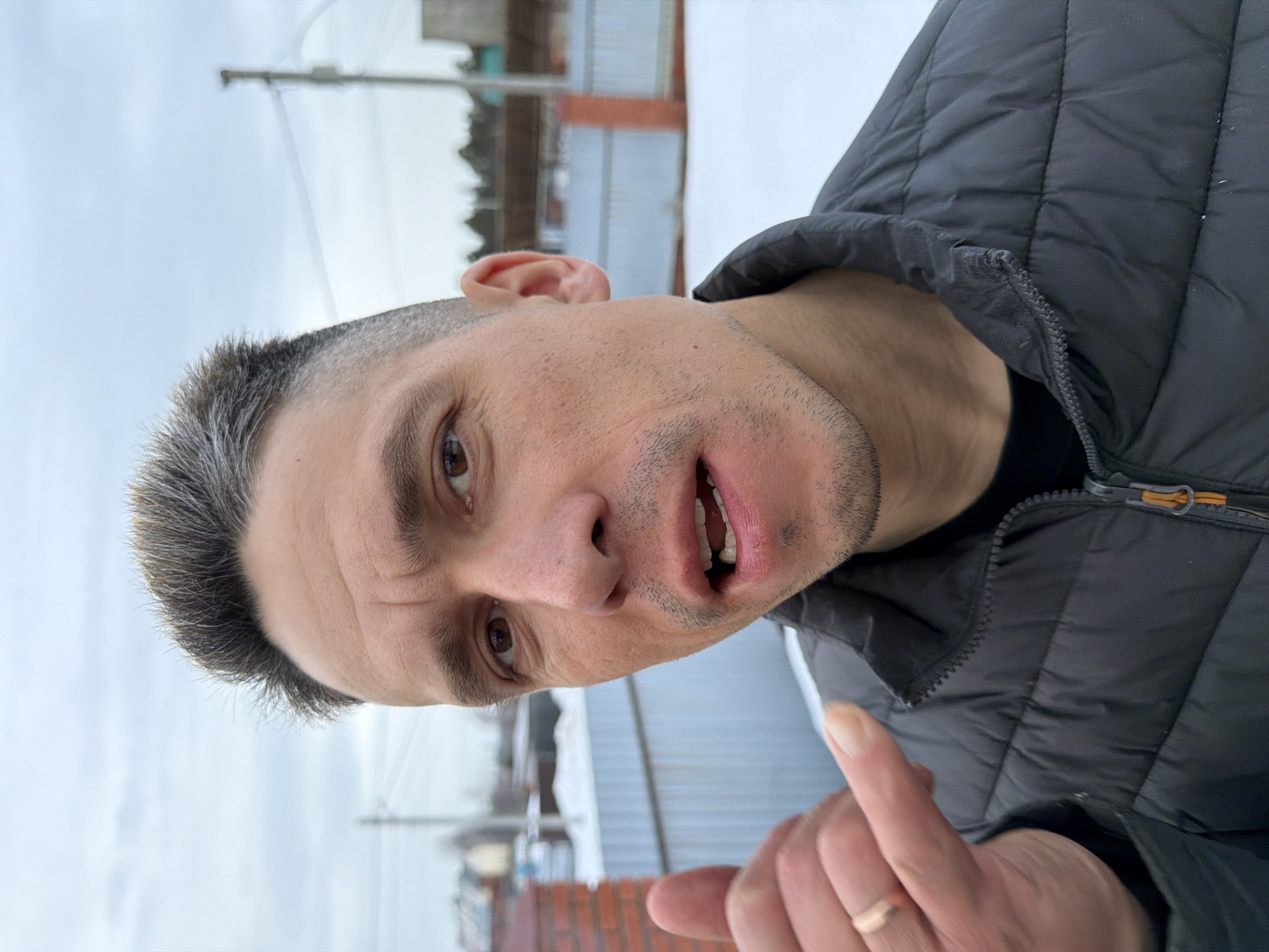 An adult male stands outdoors in a snowy environment within Chaikovsky, Russia. He is positioned slightly off-center to the right, looking directly at the camera with a partially open mouth, revealing his upper teeth. His hair is short, dark, and graying on top with faded sides. He wears a dark gray puffer jacket over a black t-shirt. His left hand is raised, performing a "call me" gesture with the thumb and pinky extended and the middle fingers folded. A gold-colored ring is visible on the fourth finger of his left hand. The background features a light gray, overcast sky. Below the sky, the ground is covered in snow. A light-colored corrugated metal fence runs horizontally across the mid-ground. To the left, a portion of a red brick fence post is visible. In the far background, blurred residential structures, utility poles, and scattered trees indicate a suburban or semi-rural setting. Another red fence post is visible behind the male's right shoulder, further along the metal fence.