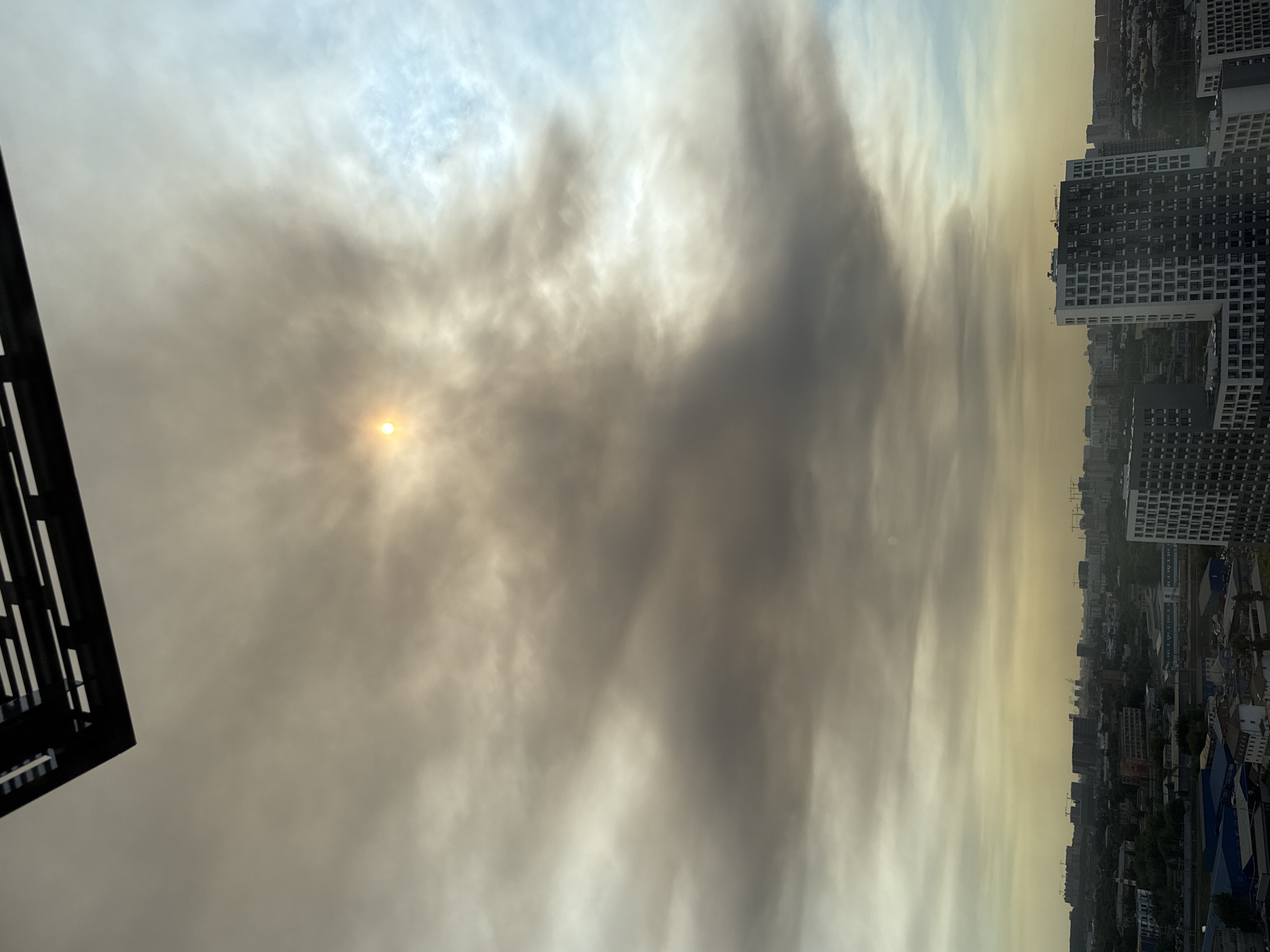 The image captures a view of the city of Lyubertsy, Russia, from an elevated position. The foreground features the corner of a dark, angular structure, possibly a building overhang or a trellis, partially obscuring the top left of the frame. Below this, the sky is filled with thick, swirling clouds, tinged with yellowish-brown hues, suggesting smoke or heavy pollution. The sun is a faint, bright circle peeking through the dense cloud cover, casting a diffused glow.
The middle ground reveals a sprawling urban landscape with numerous buildings of varying heights and architectural styles. Clusters of residential apartment buildings with many windows are prominent, some appearing modern and tall, others smaller and older. Construction cranes are visible in the distance, indicating ongoing development. Trees and greenery are interspersed between the buildings, though they appear somewhat muted by the atmospheric conditions. Power lines or industrial structures with smokestacks can also be discerned in the cityscape.
The horizon line is low in the frame, emphasizing the vastness of the sky and the layered structure of the city. The overall lighting is dim and hazy, characteristic of a day with poor air quality. There are no visible people or animals in the scene. The image conveys a sense of urban sprawl under a challenging sky.