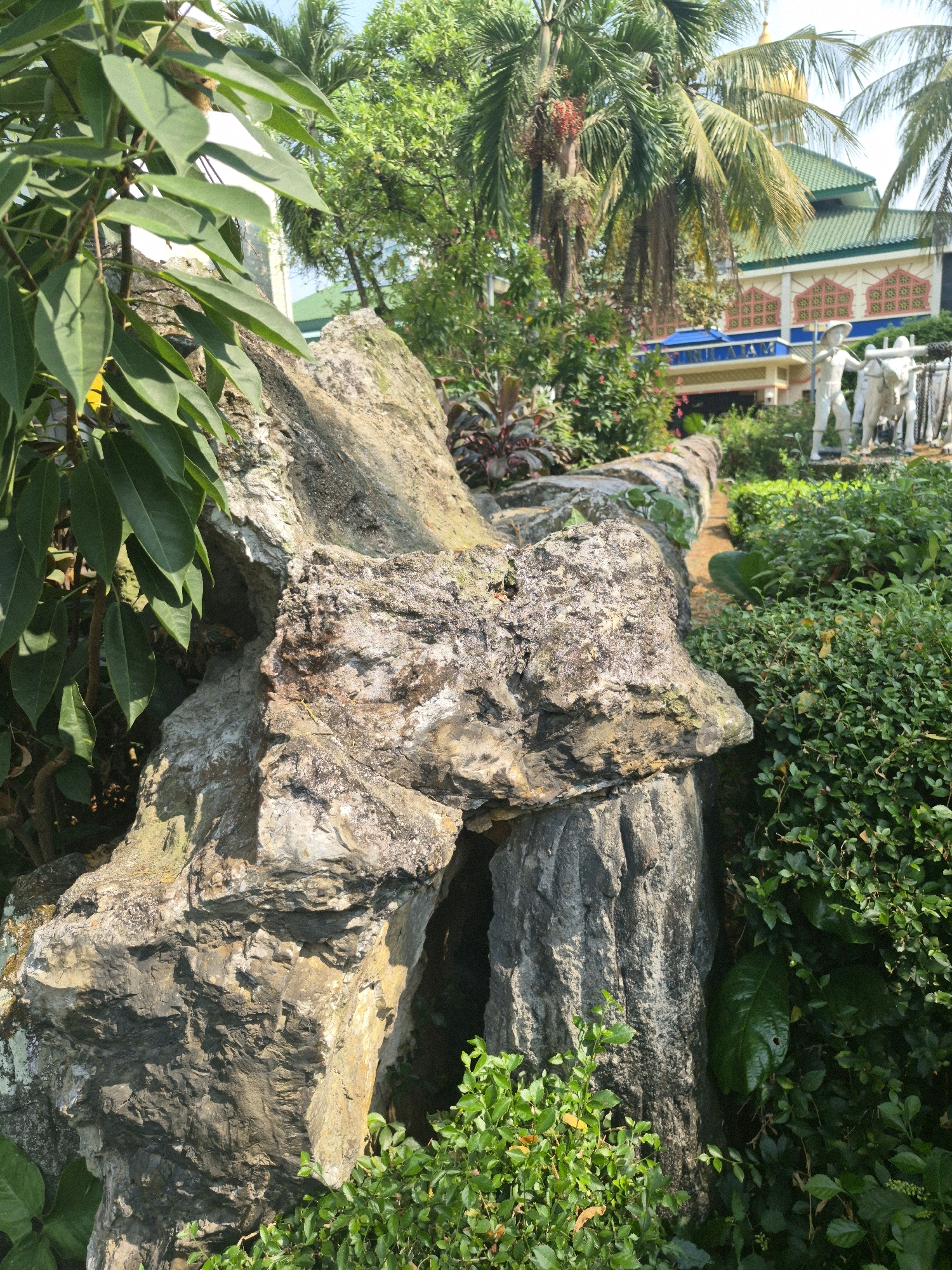 This image depicts an outdoor scene in Central Jakarta, Indonesia, with a focus on a large, weathered rock formation in the foreground. Lush green foliage surrounds the rock, with some broad leaves visible on the left and denser shrubbery on the right. Behind the rock formation, a pathway or garden bed extends into the background. Further back, several white statues are visible, depicting figures, one appearing to be a farmer with a yoke of oxen. Beyond the statues, a building with a green roof and patterned windows can be seen, partially obscured by palm trees and other vegetation. The overall impression is of a well-maintained garden or park setting, possibly a tourist attraction or cultural site within the city.