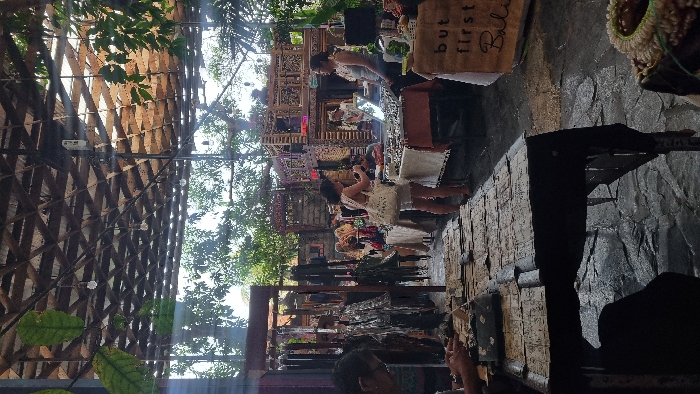 This image depicts a semi-outdoor market or craft fair located in Pecatu, Indonesia. The scene is illuminated by natural daylight filtering through a slatted wooden roof from which several bare light bulbs are suspended. Lush green foliage is visible around the perimeter and through openings in the roof structure. The ground consists of uneven, dark-toned paving stones.
In the foreground on the left, a person wearing glasses is partially visible, seated at a dark wooden table covered with numerous flat, possibly paper or textile, items. In the mid-ground, multiple market stalls are arrayed. To the left, racks display hanging items, appearing to be leather goods or textiles. Towards the center-right, a woman with dark hair, wearing light-colored shorts and a white top, stands facing a stall. She holds a mobile phone, directed towards the merchandise. She carries a white tote bag with visible text on its side.
Opposite her, behind the stall, stands a woman, likely a vendor, wearing a light-colored sleeveless top and glasses, observing the browser. The stall between them displays an array of small, reflective objects, consistent with jewelry, laid out on a light-colored cloth, with a small mirror also on the table. To the right of this stall, a cardboard sign reads "but first Bali".
In the background, an intricately carved, multi-tiered wooden structure is prominent, possibly serving as a large stall or decorative element. Other individuals are discernible in the background, moving through the market space.