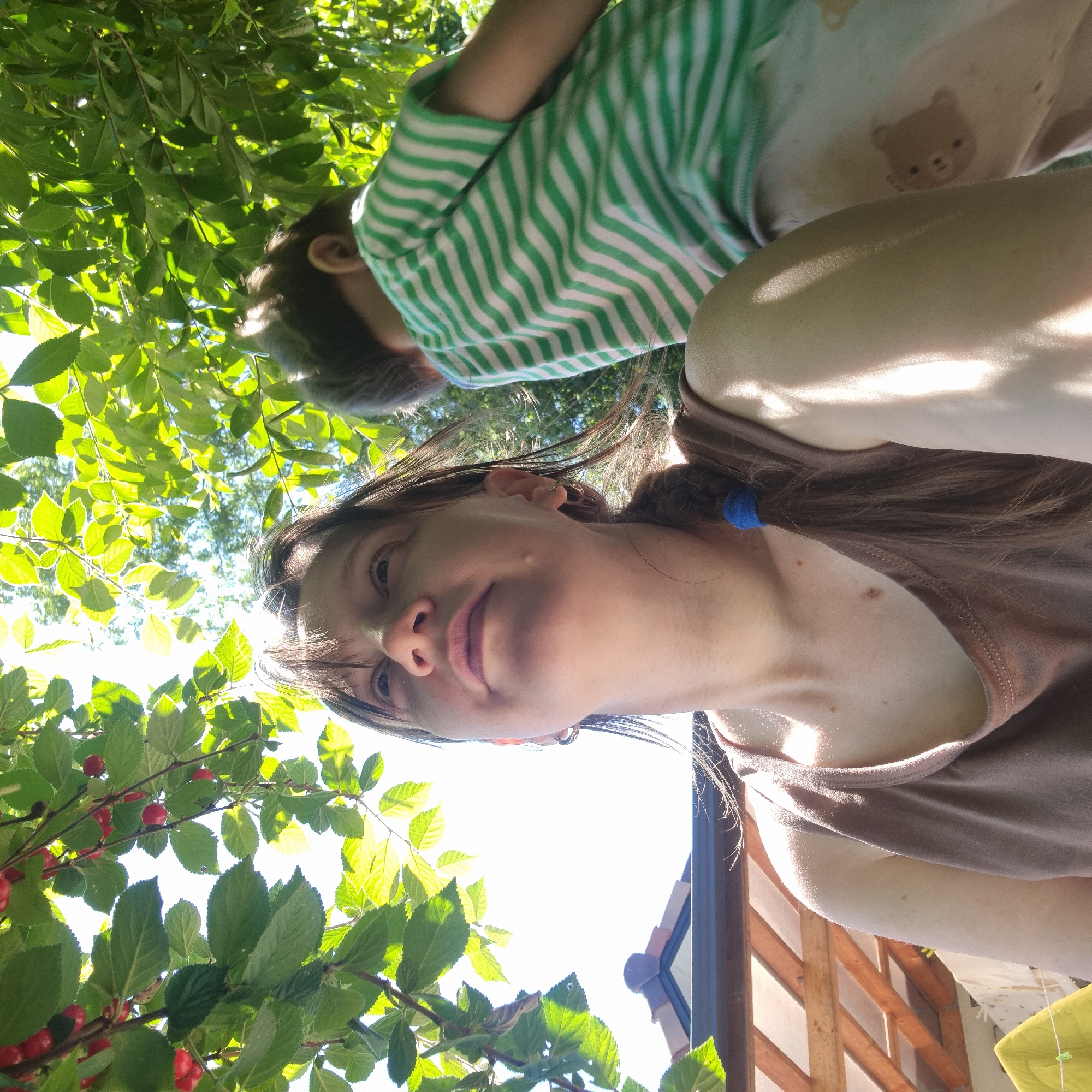 In Dunnae-myeon, South Korea, a low-angle, outdoor, close-up photograph captures two individuals beneath a canopy of green leaves and red cherries. A woman in the foreground, wearing a brown tank top, looks upwards. Her brown hair is tied back with a blue elastic band, and she has hoop earrings. Beside her, slightly out of focus and to the right, a child wearing a green and white striped t-shirt is visible. The background reveals a clear sky and a glimpse of a structure with a sloped roof and wooden beams. Sunlight filters through the leaves, creating dappled light on the subjects.