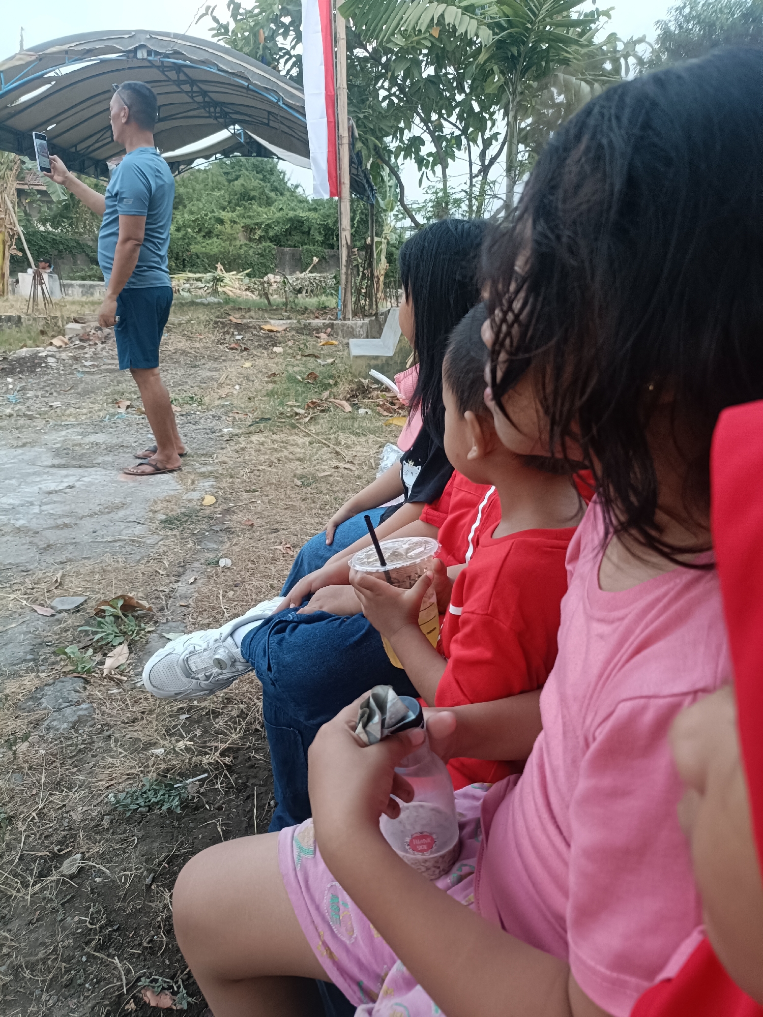 In the city of Kediri, Indonesia, a man in a blue t-shirt and shorts stands outdoors, holding a mobile phone to take a picture. He is positioned to the left of the frame. In the foreground, a group of individuals, including children, are seated on the ground. One child in a red shirt holds a plastic cup with a straw. Another child in a pink shirt is visible to the right. The environment appears to be an outdoor area with dry grass and scattered debris. A white and red flag is visible in the background, partially obscured by trees. The overall scene depicts a casual outdoor gathering.