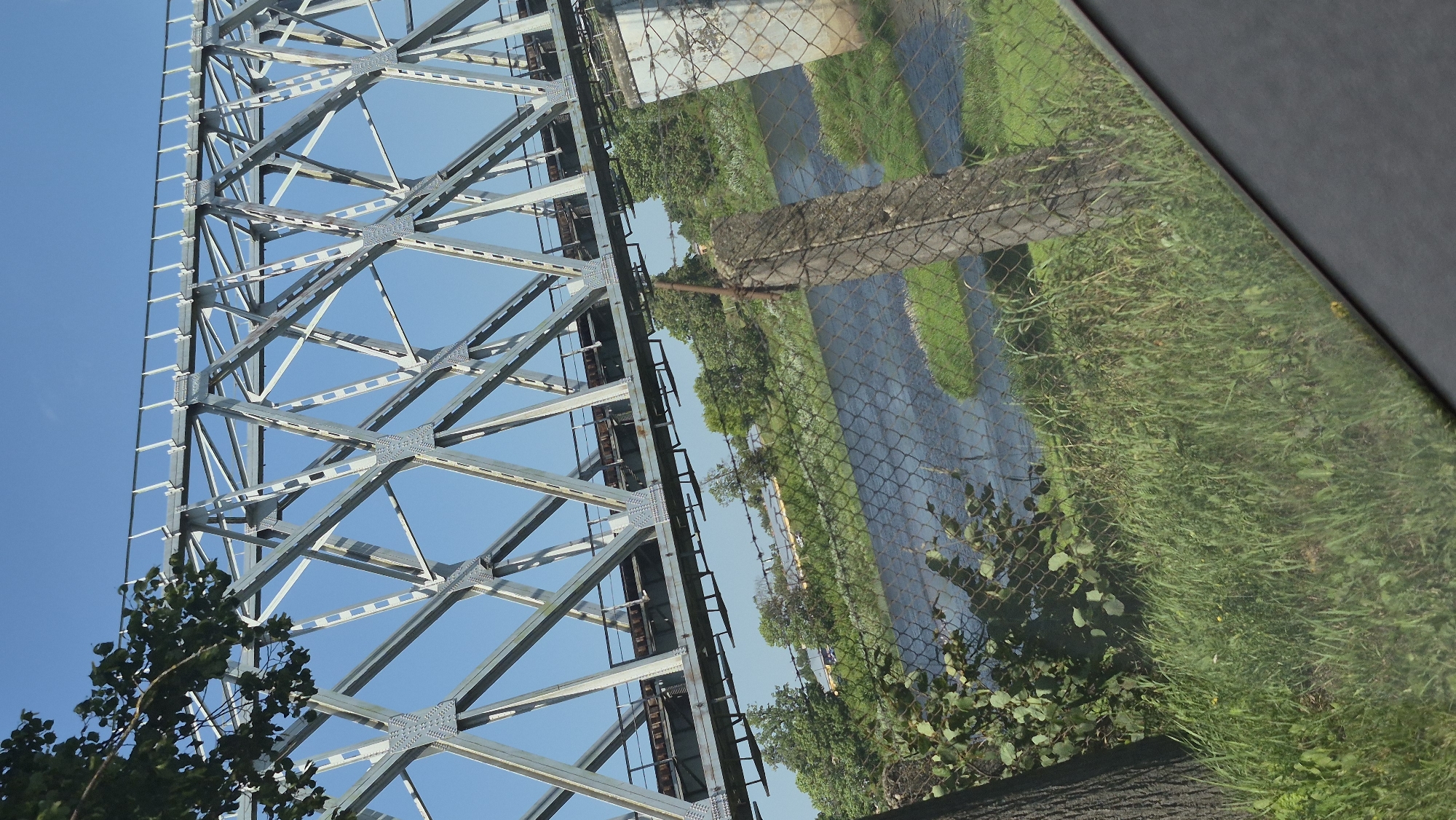The image shows a view of a metal truss railway bridge over a river. The bridge is made of interconnected steel beams forming a lattice structure. The railway tracks are visible on top of the bridge. Below the bridge, a river flows through a green, grassy landscape. There is a chain-link fence in the foreground with a wooden post. Trees and vegetation are visible on the riverbanks. The sky is clear and blue. The scene appears to be outdoors, likely in a semi-rural or industrial area, given the presence of a railway bridge and a river. The angle of the photograph suggests it was taken from a moving vehicle or elevated position looking down at an angle.
