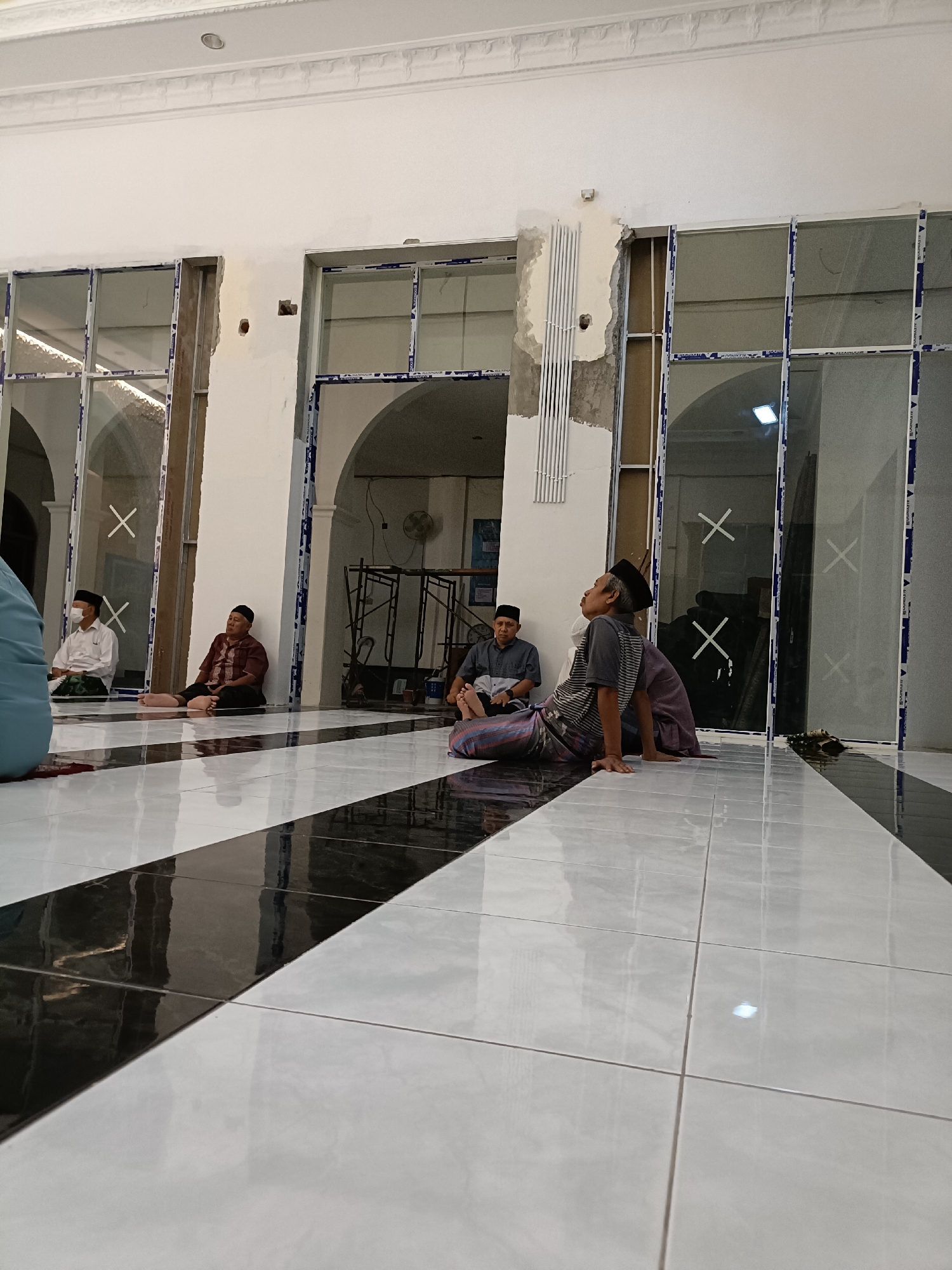 The image shows a partially constructed or renovated interior space, likely a mosque, in Kediri City, Indonesia. The floor is tiled with large, glossy white and black tiles arranged in a diagonal pattern. Several men are seated on the floor, some appear to be praying or in quiet contemplation.
On the left, a man in a white shirt and black peci, wearing a surgical mask, sits with his legs crossed. Next to him, a man in a maroon shirt and black peci is seated with his legs extended. Further to the right, a man in a grey shirt and black peci sits with his legs extended, looking upwards. Another person, partially visible, is seated to his right, wearing a purple garment.
The background features large glass windows with white frames, some of which are open. The walls are white, with visible construction elements such as exposed wiring and unfinished plaster around the window frames. Architectural features like arches and decorative molding are also present. In the background, scaffolding and what appears to be a ventilation fan are visible. The lighting suggests an indoor setting, possibly during daytime.