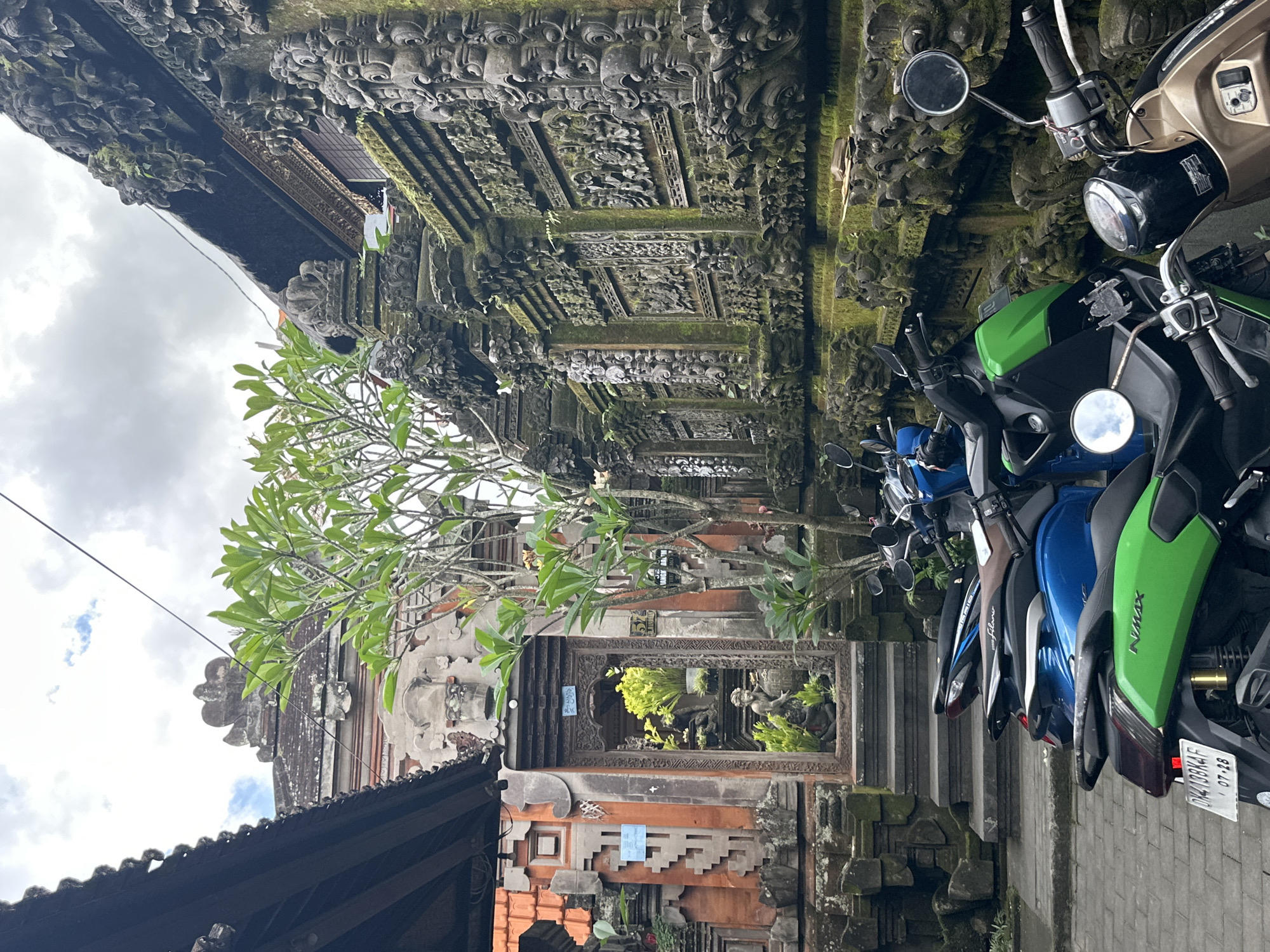 The image depicts a street scene in the Ubud District, Indonesia, likely in front of a traditional Balinese building. Several scooters are parked along a stone pathway in the foreground. The scooters are of various colors, including green, blue, and brown. Behind the scooters, a pathway leads up to a ornate, carved wooden doorway, framed by red brick walls with intricate stone carvings. A small statue is visible through the doorway. The buildings feature traditional Balinese architecture with moss-covered stone carvings and decorative elements. A tree with green leaves stands near the center of the frame. The sky above is cloudy, suggesting overcast weather. The overall atmosphere is one of cultural immersion and daily life in a Balinese setting.
