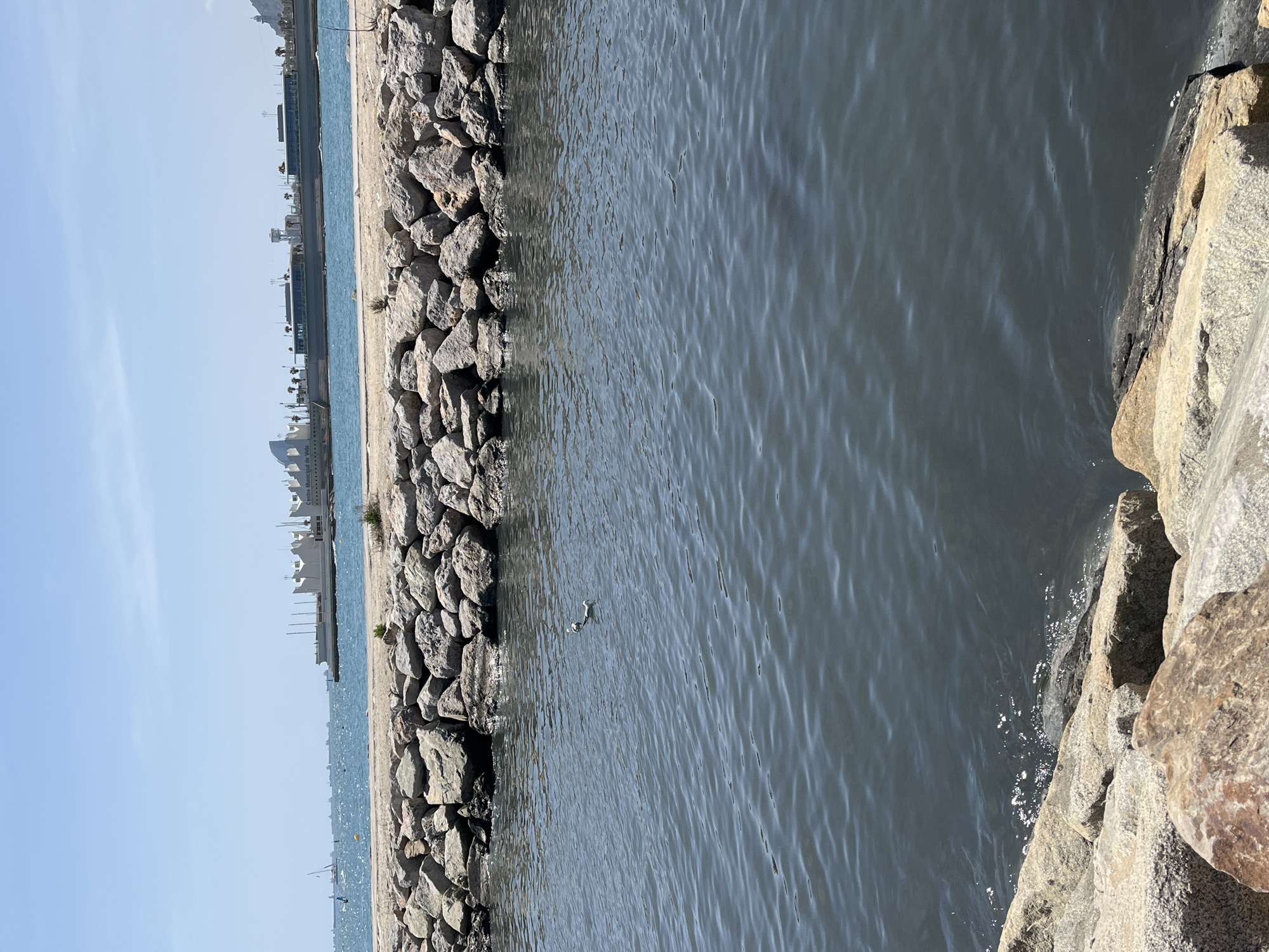 This image depicts a body of water bordered by a stone breakwater, likely the Mediterranean Sea along the Barcelona coastline. The water is calm with visible surface ripples. The breakwater is constructed from large, irregularly shaped gray rocks. Beyond the breakwater, there's a sandy beach and a series of low-rise buildings and structures, potentially part of the Barcelona harbor or port area. The sky is clear and blue. The image appears to be taken during daylight.