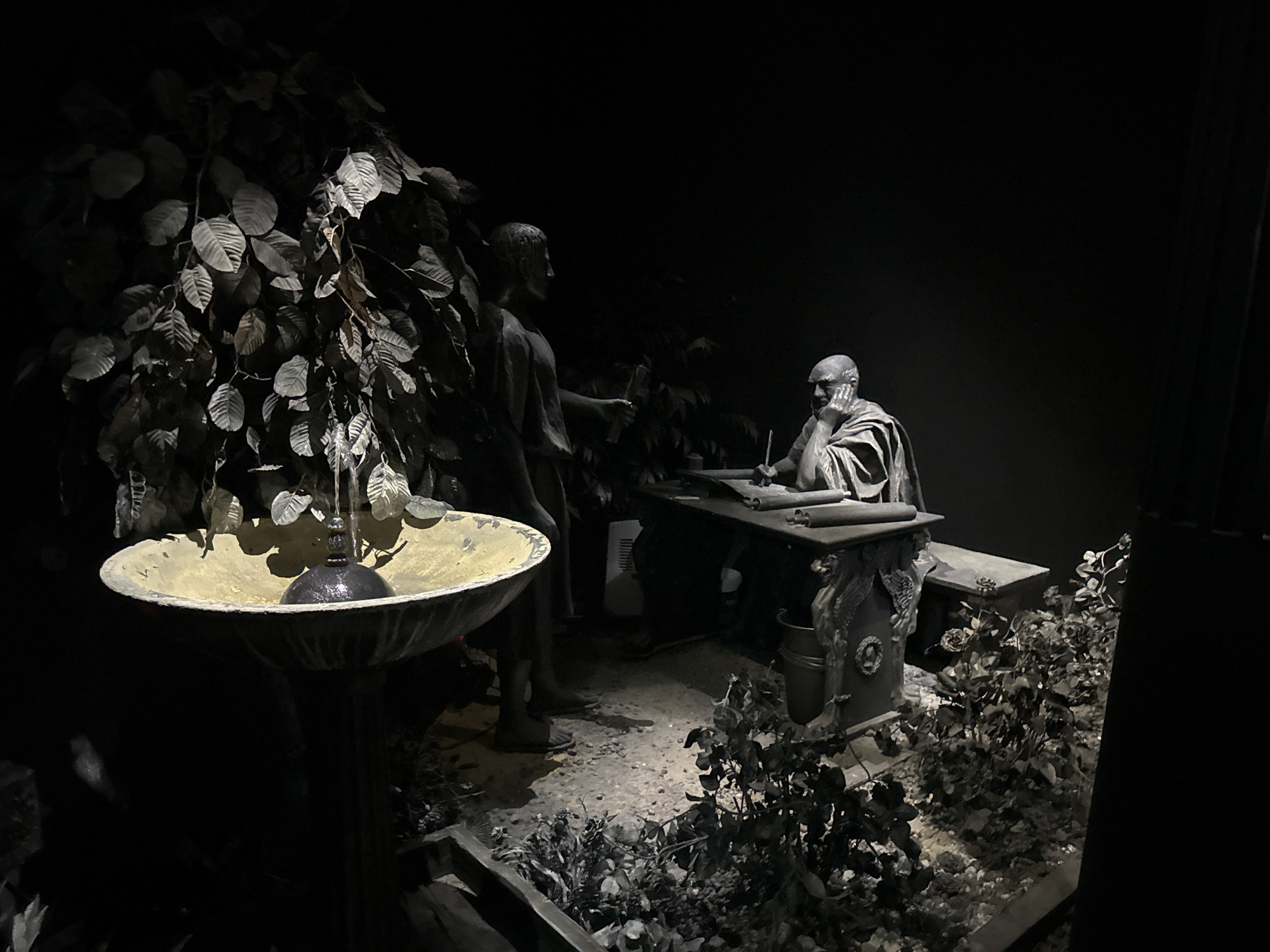 The image depicts a dark, theatrical scene featuring two statues. In the foreground left, a birdbath-like structure with a decorative rim and a central hanging element is visible, partially obscured by dark foliage. To the right of the birdbath, two statues are positioned. One statue, standing and facing towards a seated figure, appears to be holding a tool or object. The second statue, seated at a desk, is depicted with a hand resting on its chin, appearing contemplative or engaged in writing, with a pen or stylus in its other hand. The seated figure is wearing a draped garment. The setting includes a dark background, suggestive of an indoor exhibit or a dramatized outdoor setting, with scattered gravel and more dark foliage around the base of the statues. The lighting is dramatic, casting strong shadows and highlighting specific elements of the scene. There is no direct indication of the city of Tamar Regional Council, Israel within the image itself.
