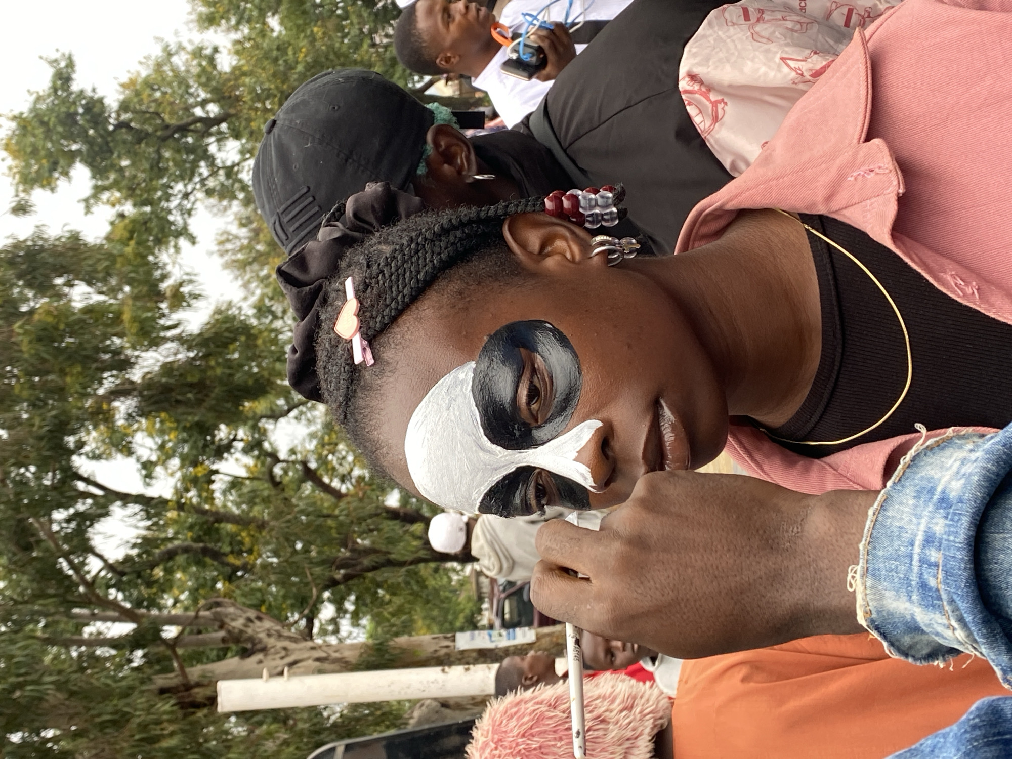 In Bar Jirgi Summa, Nigeria, a young person with dark skin is having their face painted with a black and white design resembling a panda or skull. The painting is in progress, with a brush held by the subject's left hand nearing their eye. They are wearing a pink denim jacket over a black shirt and a thin gold chain. Their hair is styled in braids, adorned with a pink clip and beaded extensions. Other people are visible in the background, including a person in an orange top to the left and individuals in darker clothing behind the subject. The environment appears to be outdoors, with trees and an overcast sky.