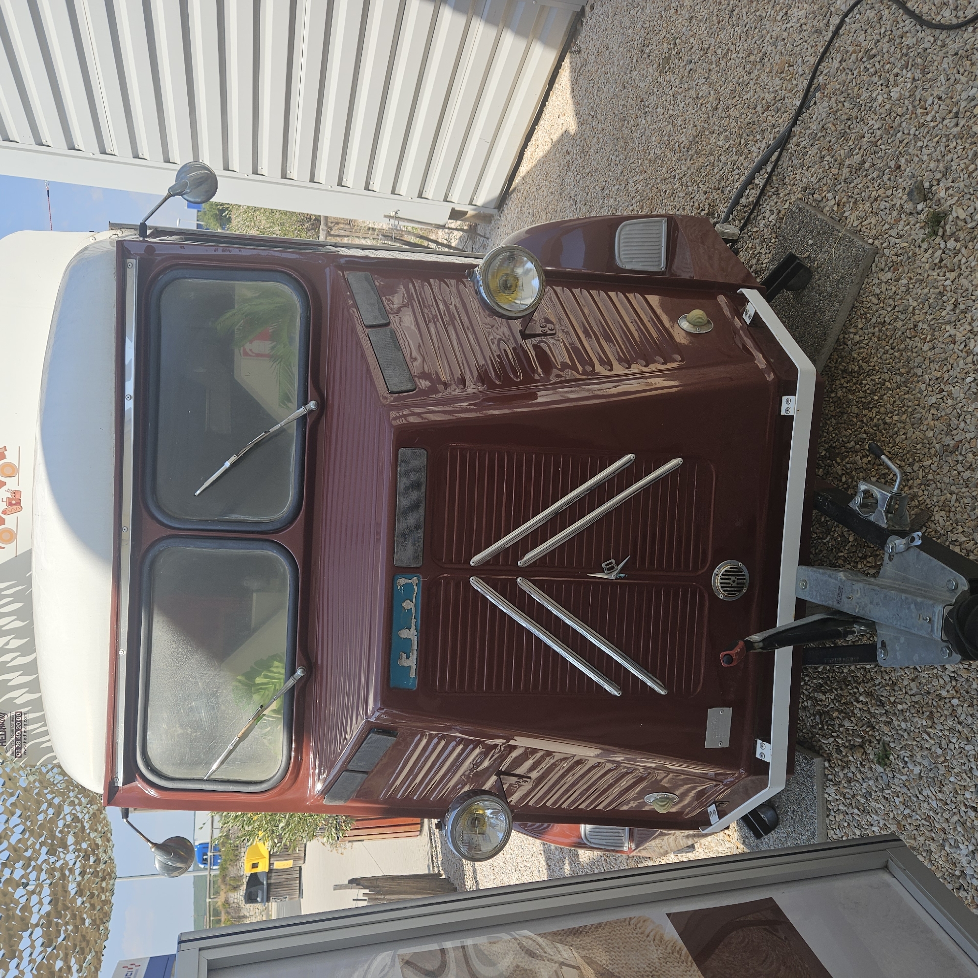 The image shows the front of a vintage, maroon-colored vehicle, possibly a food truck or camper, parked on a gravel surface. The vehicle has a distinctive ribbed front panel and two large windshields. Round headlights are mounted on either side of the front. A tow hitch is visible at the bottom. In the background, there is a white corrugated metal building, some vegetation, and a camouflage-patterned shade structure overhead. A blue and yellow trash bin is also visible in the mid-ground. The scene appears to be outdoors in a semi-industrial or commercial area. The image is taken from a frontal perspective, looking slightly upwards at the vehicle. There are no people visible in the image. The location is identified as the city of La Palme, France, suggesting it is a public space within that area.