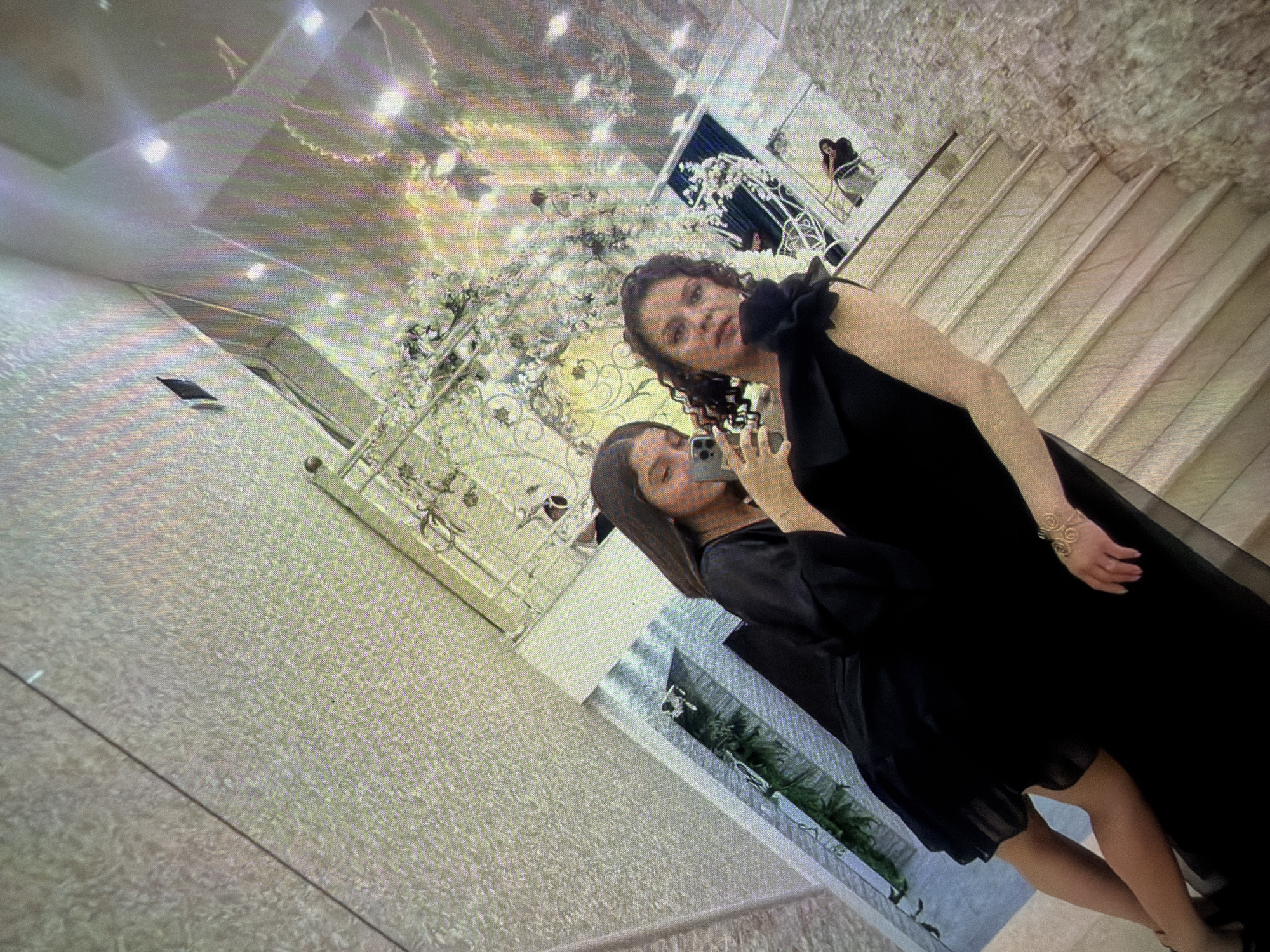 Two women are visible in a dimly lit room of Unknown, Unknown, near a decorated staircase. The woman closer to the camera is holding a cell phone, presumably taking a selfie. Both women are dressed in dark-colored clothing. The environment includes ornamental ironwork, white floral decorations, and stairs covered in a light-colored material. The scene is angled and appears to be a photograph of a monitor screen.