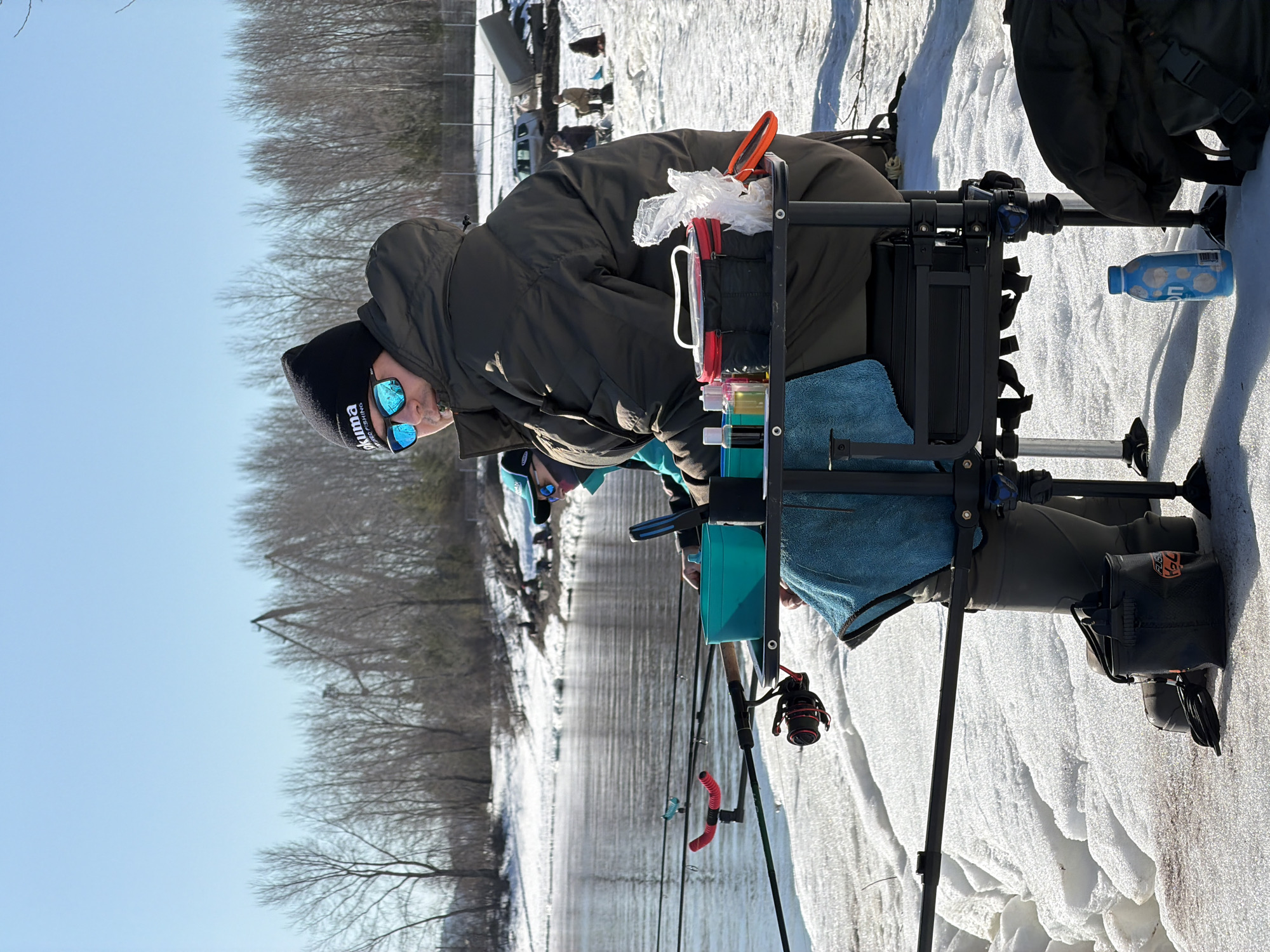 A man is fishing on a snow-covered river in Chaikovsky, Russia. He is wearing a dark green coat, a black hat, and blue mirrored sunglasses. He is sitting on a black fishing platform, with a fishing rod in the water. Other people are visible in the background, also fishing. The river is partially frozen, and bare trees line the riverbank. The sky is clear and blue. Snow covers the ground.