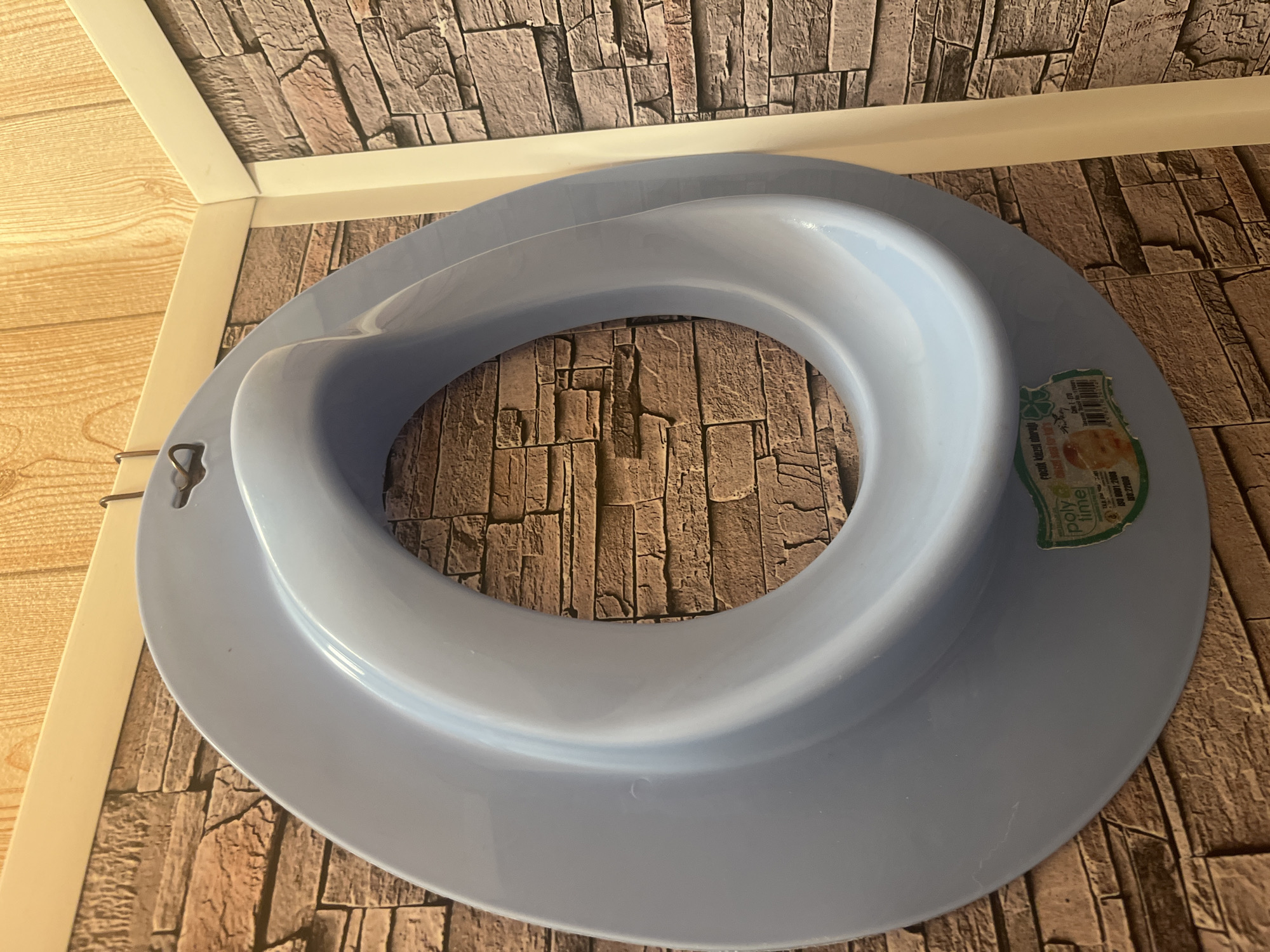 A light blue plastic toilet seat trainer for children is hanging on a hook against a wall with a faux brick pattern wallpaper. The trainer has an oval opening and a wider base. A small sticker with product information and a picture of a baby is attached to the lower right side of the trainer. The wall also has a section of light wood-patterned wallpaper at the top. The image is taken from a medium distance, focusing on the toilet seat trainer. This scene is likely in a residential bathroom in Burgas, Bulgaria.