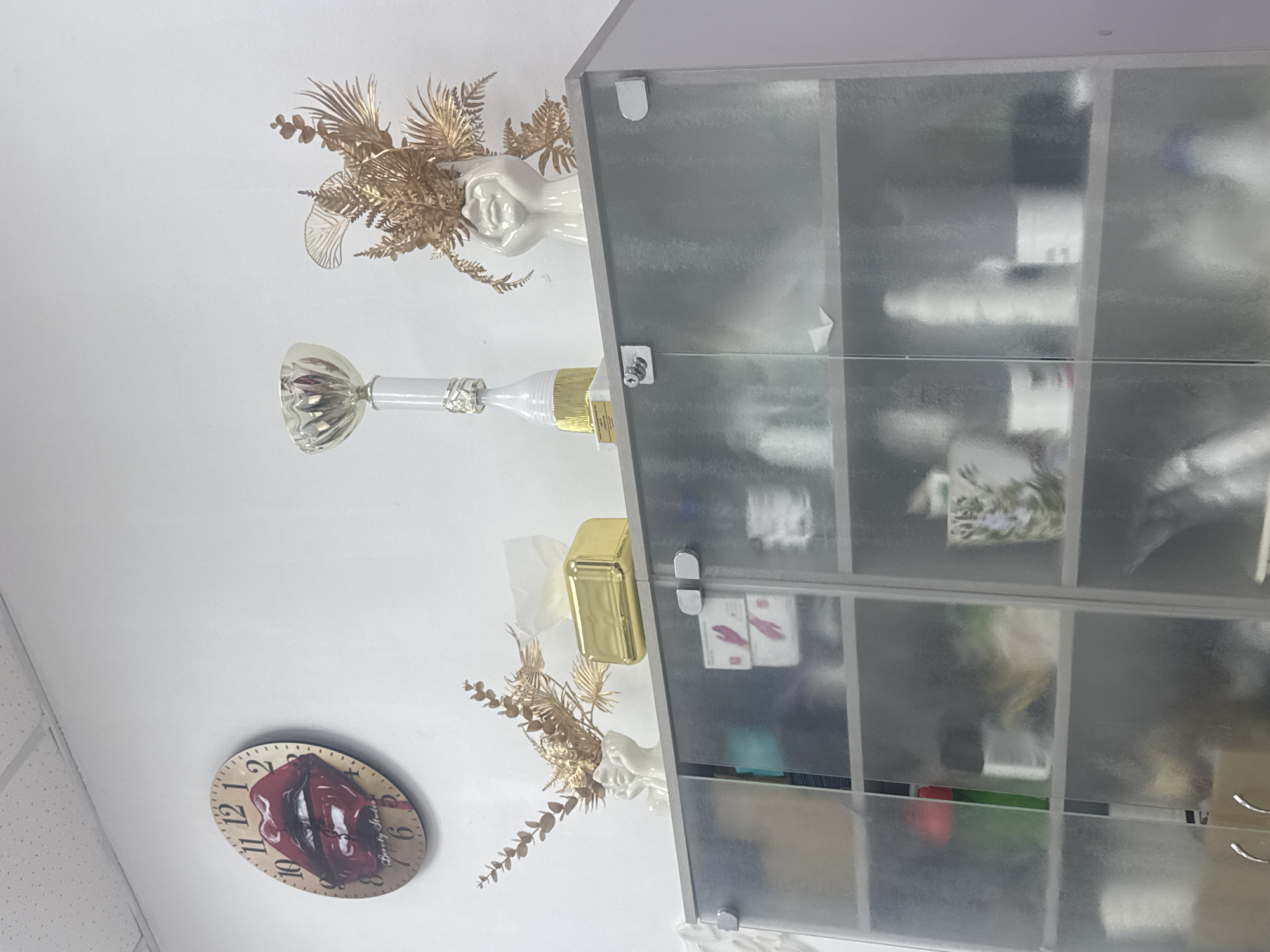 The image shows a close-up of a frosted glass-fronted cabinet in what appears to be a beauty salon or spa, likely located in Komsomolsk-on-Amur, Russia. The cabinet has two doors, each with two shelves, revealing various bottles, jars, and other products. Above the cabinet, on the wall, is a round clock with a lip design and the words "Beauty Studio." To the right of the clock, there are decorative golden foliage in a white vase and a golden tissue box. A trophy is also visible on top of the cabinet. The overall scene suggests a professional setting focused on beauty treatments.