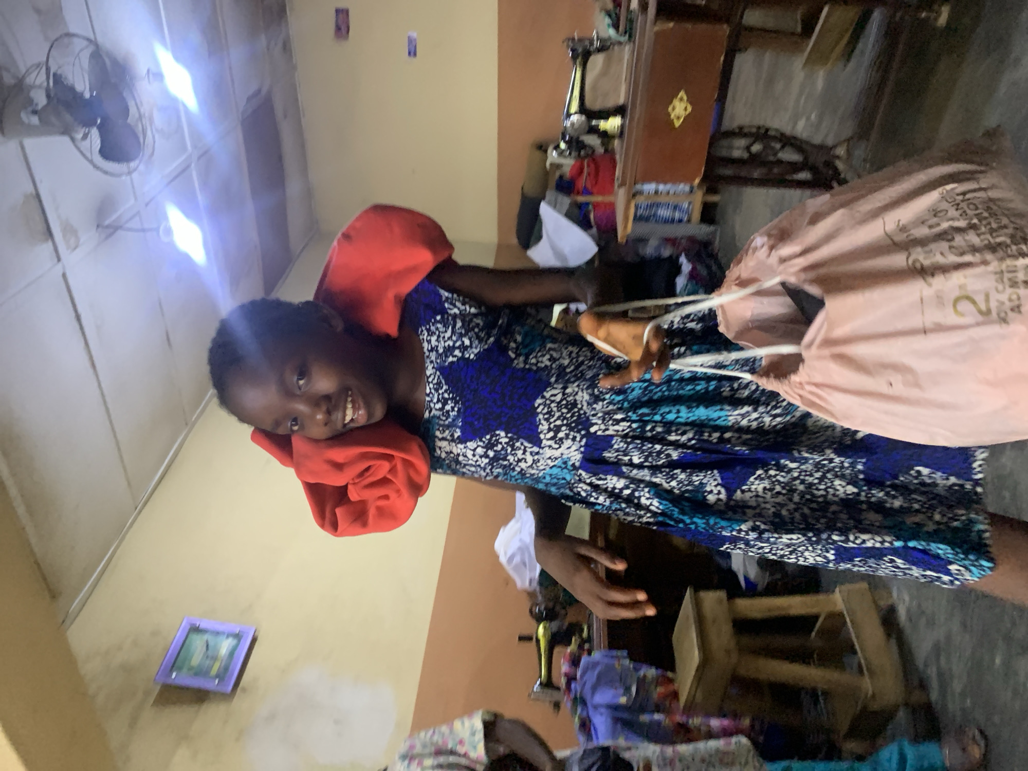 A young Black woman is standing in what appears to be a small sewing workshop in Jos, Nigeria. She is wearing a colorful blue and white patterned dress, a red headscarf, and holding a large tan-colored bag. The bag has the number "2" printed on it, along with other text in smaller print. The woman is smiling and looking directly at the camera.
In the background, there are several sewing machines, pieces of fabric, and wooden stools. The walls are painted a combination of beige and light brown. A ceiling fan is visible in the upper left corner of the frame. A small framed picture hangs on the wall.