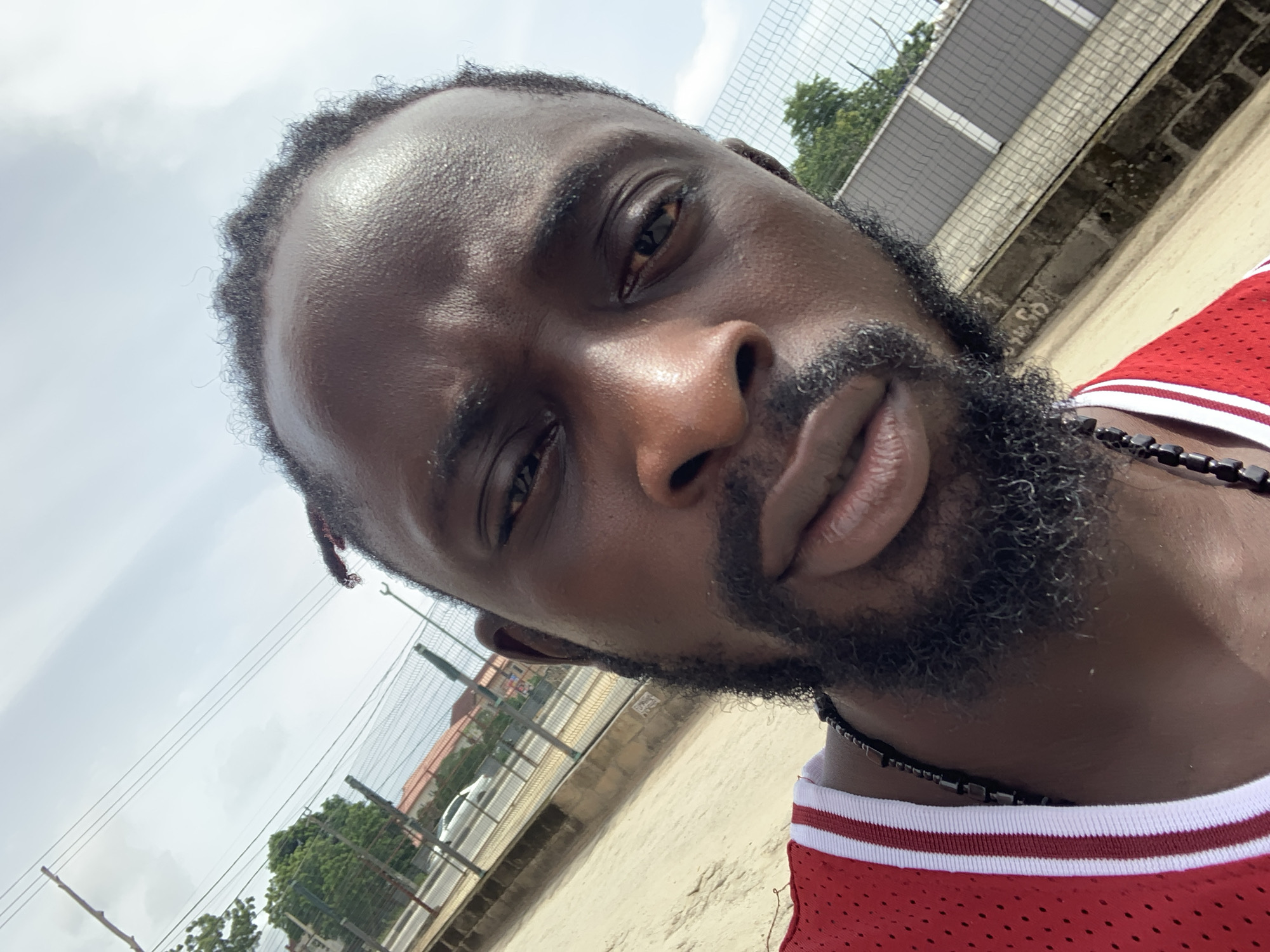 A close-up, angled selfie-style portrait features a Black man outdoors. He occupies the majority of the frame, looking directly towards the camera with a neutral to slightly pensive expression. His skin, particularly on his forehead, nose, and cheekbones, glistens, suggesting warmth or humidity. He has short, dark, tightly curled hair and a full, dark beard. He wears a red sleeveless jersey with white striped trim on the collar, and a dark, beaded necklace around his neck.
The background is an outdoor setting, likely under an overcast but bright sky during the daytime. Behind him, a chain-link fence is visible, partially obscuring what appears to be a light-colored building wall and some distant green trees. A glimpse of a white car can be seen beyond the fence. The ground in the foreground and mid-ground appears to be dirt or sand. The overall scene suggests an urban or semi-urban environment in Sangotedo, Nigeria. There is no visible text.
