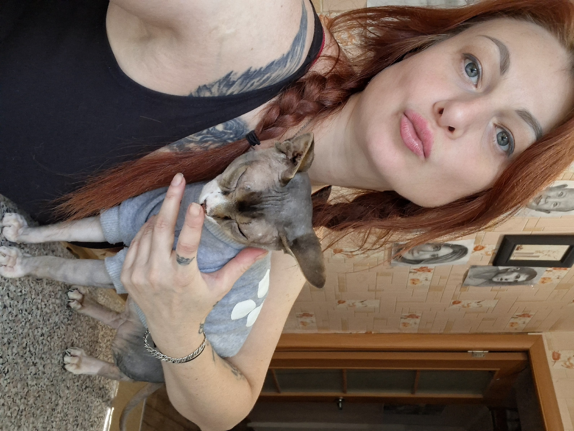 A woman with auburn hair styled in a braid and wearing a black tank top is holding a Sphynx cat, which is wearing a light blue sweater with white petal-shaped designs. The woman is making a kiss face and looking at the camera. She has tattoos on her left arm and shoulder, and a bracelet on her right wrist. The cat is facing away from the camera, with its eyes closed. They are standing on a carpeted floor, with a table visible in the foreground. In the background, there are framed pictures on a wallpapered wall, and a wooden doorframe. The overall scene appears to be indoors, likely in a residential setting. There is no information available to identify the specific location in the city of Myadzyel, Belarus.