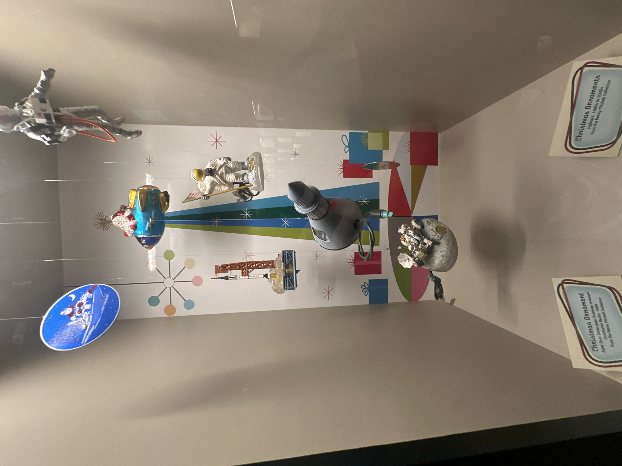 This image depicts a display case at the U.S. Space & Rocket Center in Huntsville, Alabama. The display features a collection of space-themed Christmas ornaments suspended from the ceiling. Among the ornaments are a vintage-style Santa Claus on a rocket, an astronaut in a spacesuit, a model of a Saturn V rocket launch, and a lunar module with astronauts. The background of the display is decorated with mid-century modern atomic-style graphics and gift boxes, evoking a retro-futuristic aesthetic. Two informational placards are visible at the bottom of the display, identifying some of the ornaments. The lighting suggests an indoor environment, likely a museum or exhibition space.