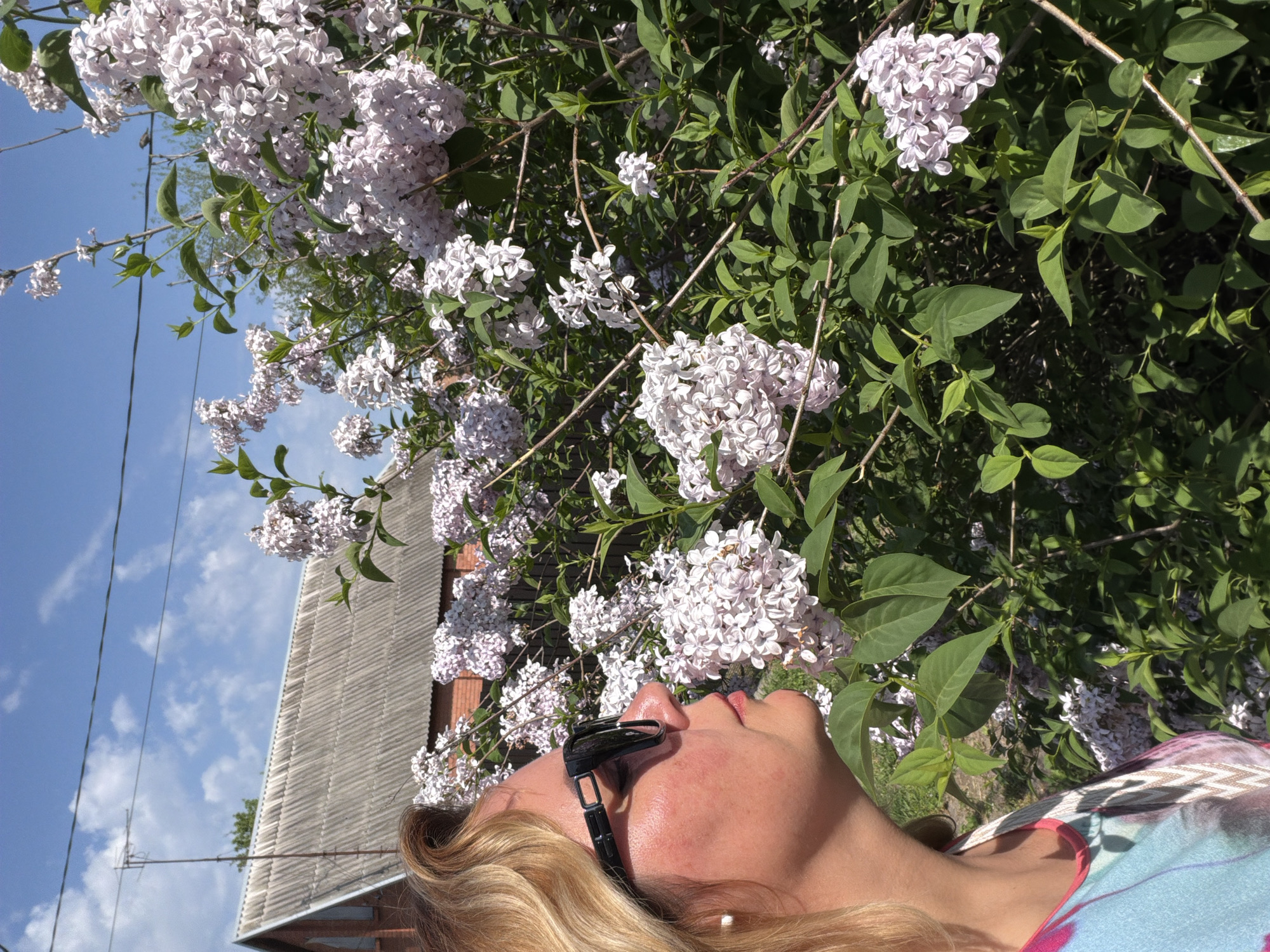 A low-angle medium shot captures a fair-skinned woman with blonde hair and sunglasses, looking upwards towards a cluster of lilac bushes with light purple and white blossoms. The woman is wearing a colorful patterned top. Behind the bushes is a blue sky with scattered clouds and a glimpse of a building with a gray, pitched roof. The location is Leningradskaya, Russia.