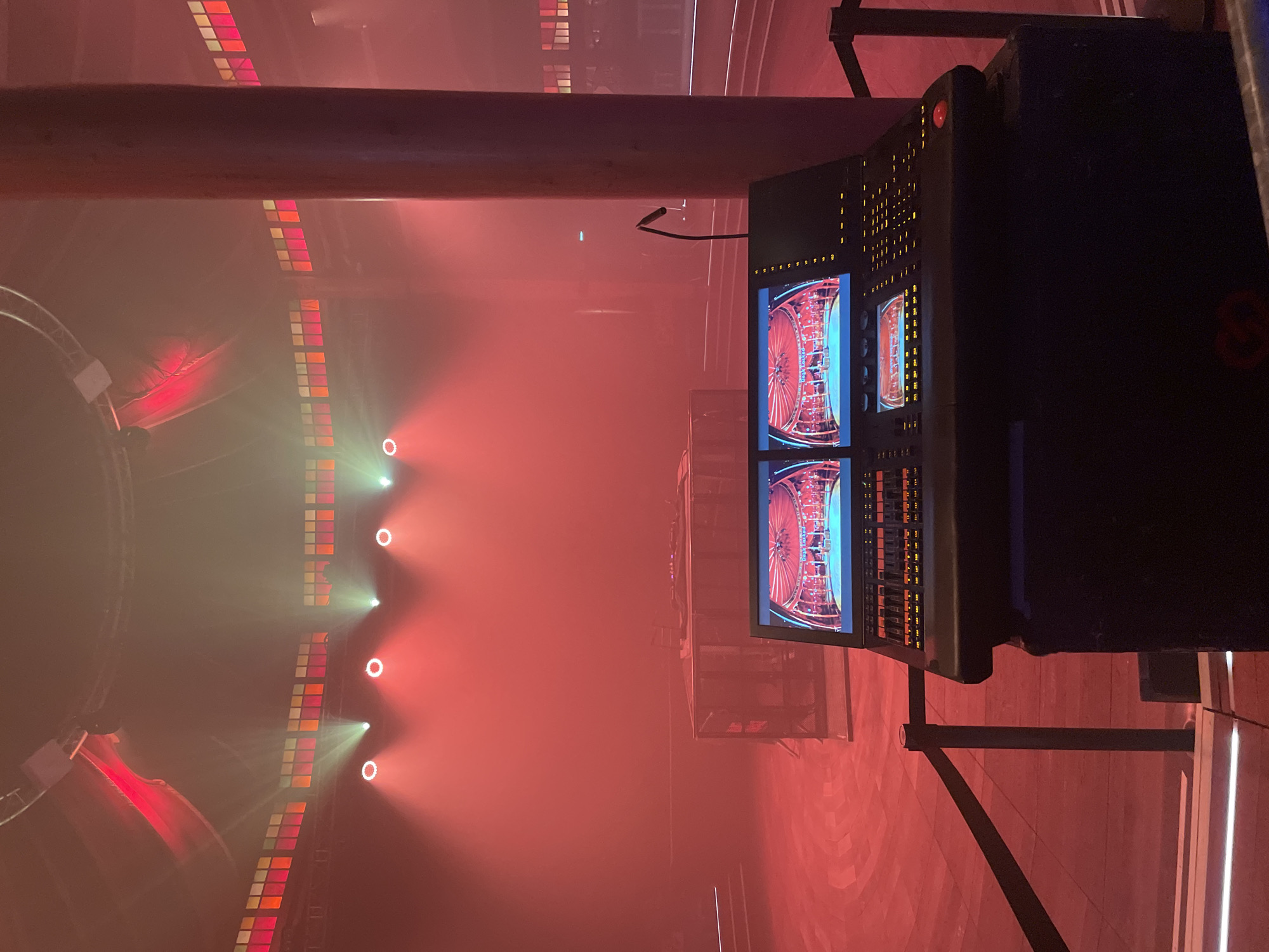 The image depicts the interior of the Cirque d'Hiver Bouglione in Paris, France, from an elevated control position. The scene is immersed in dense red atmospheric haze, illuminated primarily by red lighting.
In the immediate foreground, a black production control console is visible, featuring dual digital screens, numerous illuminated buttons, sliders, and faders. The left screen displays an interior stage set with the text "Das Glücksrad" (German for "Wheel of Fortune") visible. To the right of the console, a large, dark red cylindrical support pillar stands vertically. A black velvet rope barrier is present directly in front of the console.
In the midground, a central stage or platform constructed from metal trussing is positioned on a circular wooden floor, which exhibits a concentric pattern and is bathed in red light. Above, encircling the upper wall of the venue, a series of multi-paneled windows are illuminated in shades of red, orange, and yellow. Multiple spotlights project focused beams of white and red light through the haze towards the central stage area. No individuals are present in the frame. The overall environment suggests an event setup or rehearsal within a historic performance venue.