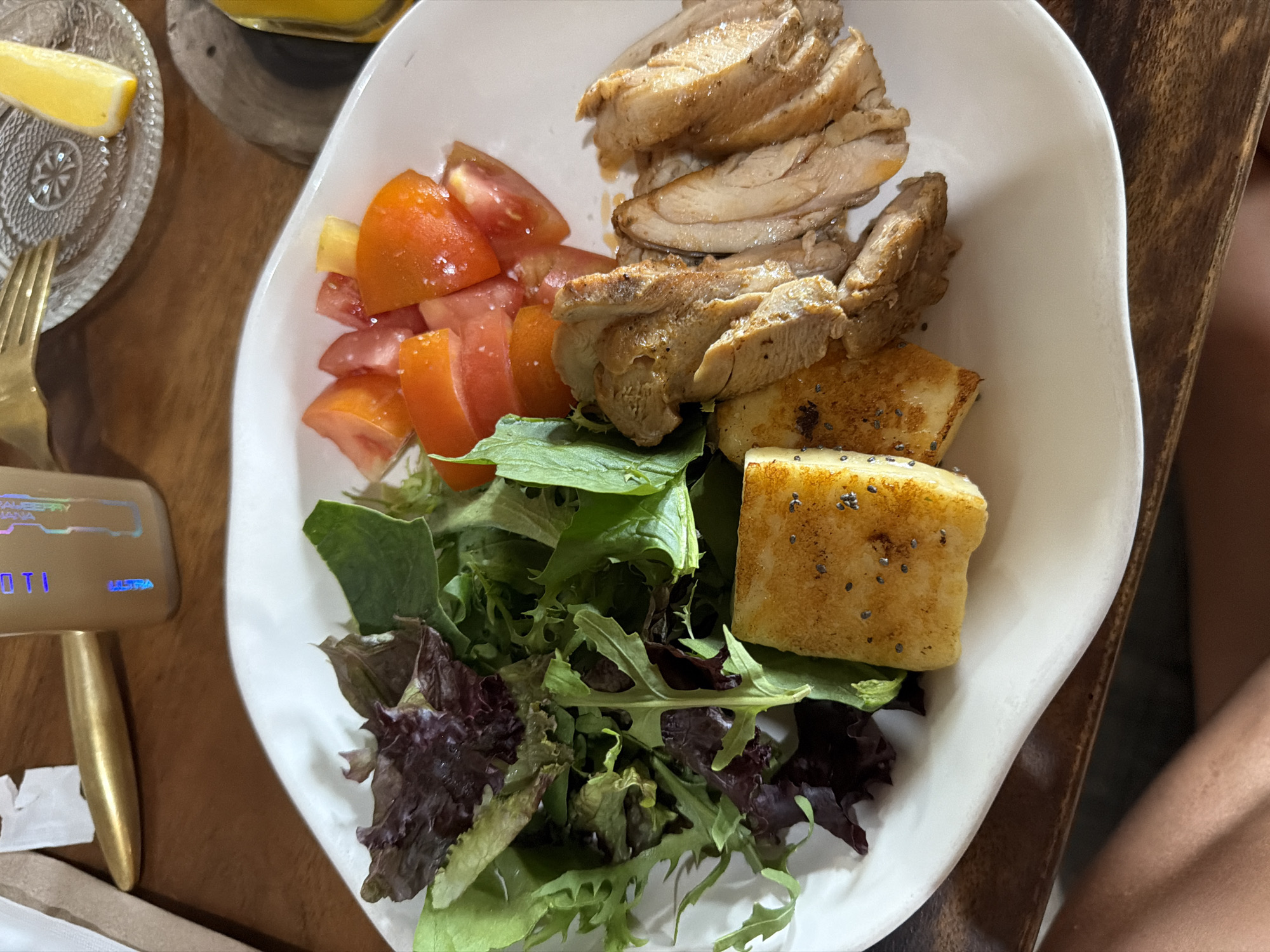 A top-down view displays a meal served on an irregularly shaped white plate, positioned on a dark brown wooden table in Canggu, Indonesia. The plate holds sliced, cooked poultry or meat on the right, alongside diced red tomatoes showing specks of white crystals, likely salt. To the left are mixed salad greens, featuring both light and dark varieties, and two rectangular, golden-brown pieces of food, possibly grilled halloumi or tofu, speckled with dark chia seeds. In the upper left background, a golden utensil handle rests near a brown paper bag. In the upper right background, a clear glass plate with an embossed starburst pattern supports a yellow lemon wedge, with a partially visible beverage bottle behind it. A portion of a person's bare leg is visible in the lower frame, indicating a diner's perspective.