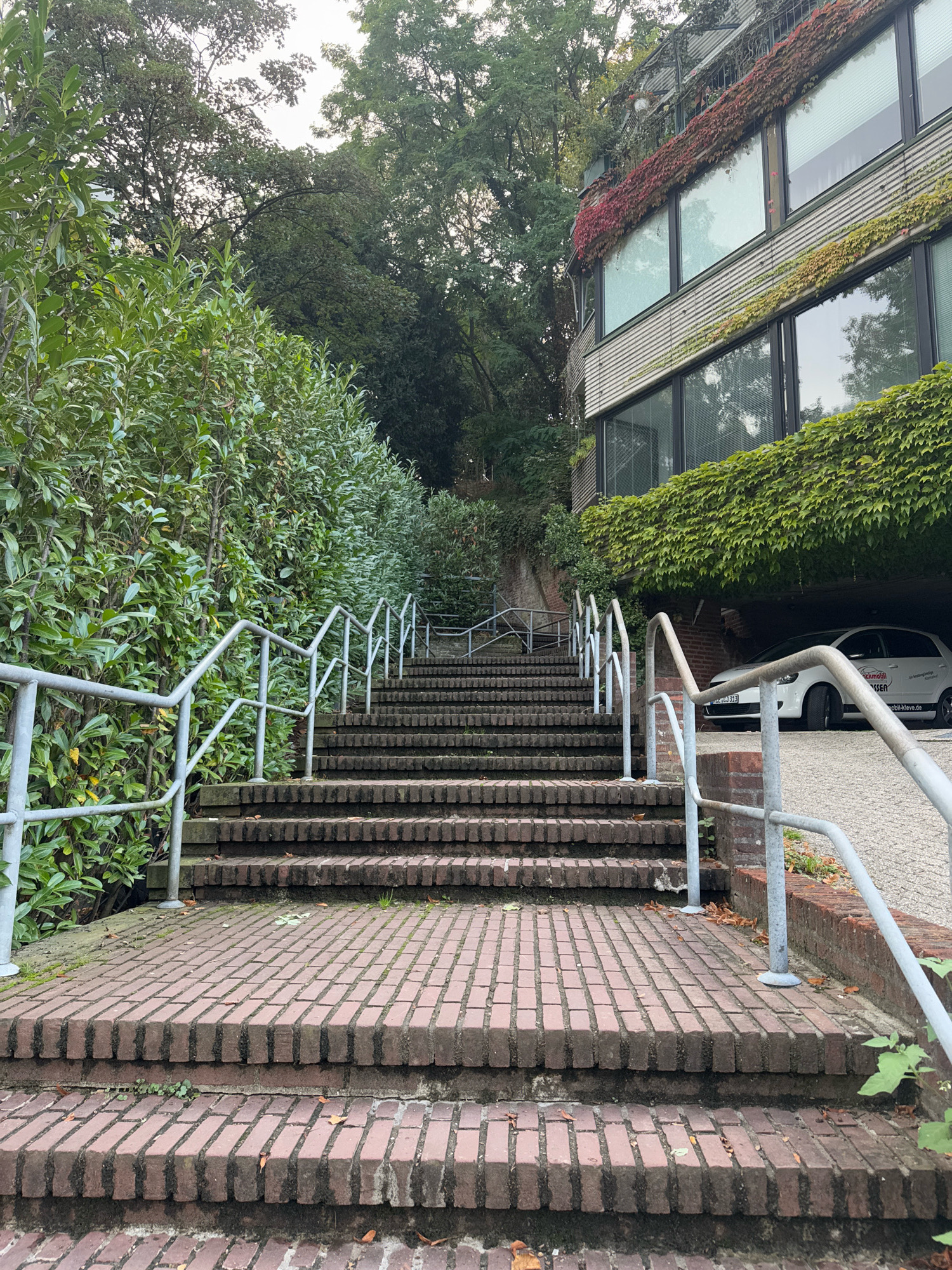 The image depicts a brick staircase with a metal railing leading upwards. Lush green bushes line the left side of the staircase, while a modern building covered in ivy is visible on the right. A white car is parked in a covered area to the right of the staircase, beneath the building. The scene appears to be an outdoor setting, possibly in a residential or commercial area. The overall impression is one of a well-maintained outdoor space with a blend of natural and architectural elements. The car has license plate KILJ 313.
