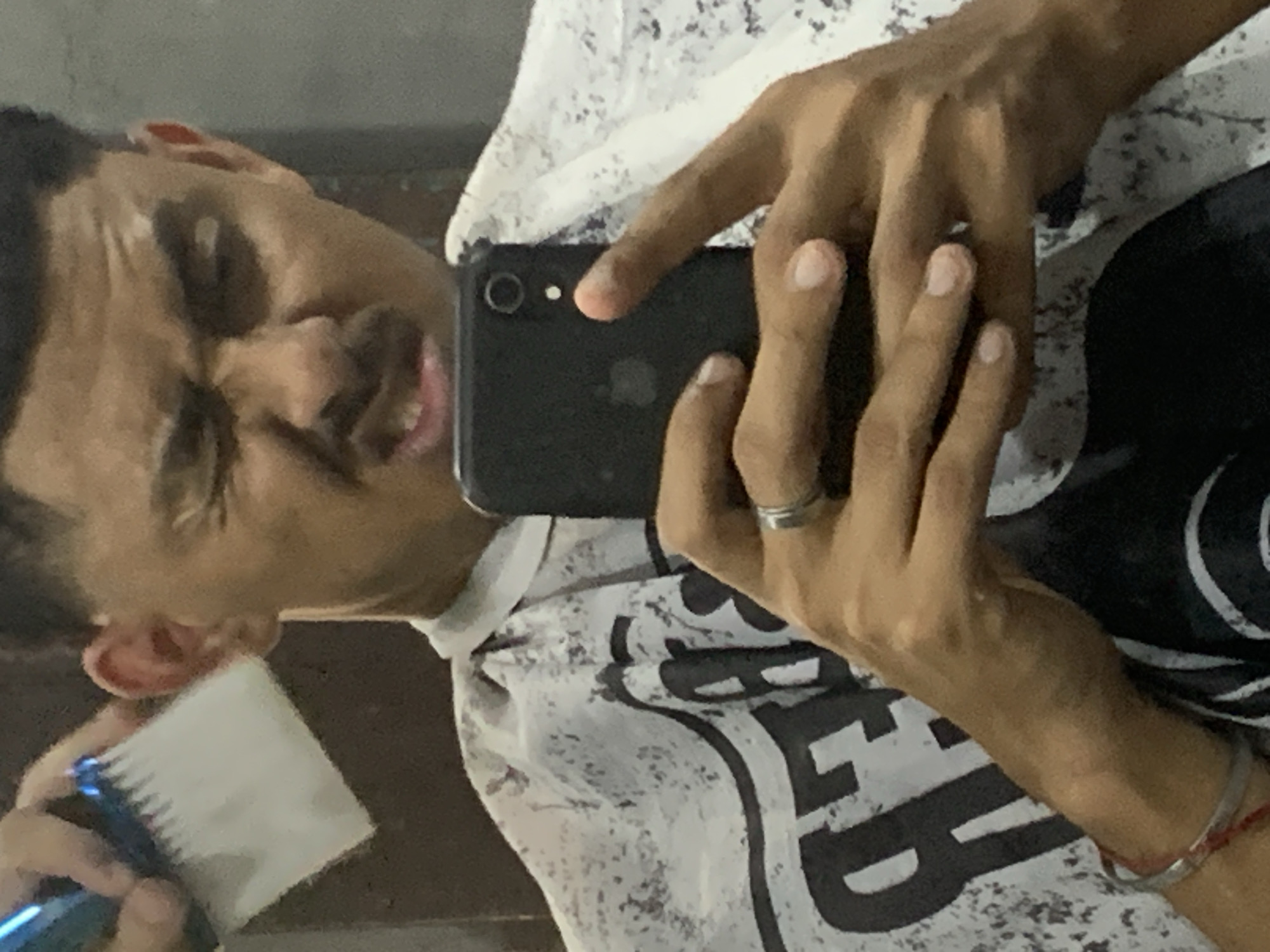 The image, taken in La Tagua, Venezuela, is a mirror selfie featuring a young man with a medium brown complexion. He is seen from the chest up, with his face turned slightly and his eyes squinting or partially closed, revealing a slight smile or smirk. He has short, dark hair.
The man holds a black smartphone, likely an iPhone, in his right hand, which reflects his face. A silver ring is visible on his right ring finger. In his left hand, he holds a blue electric hair clipper with a white comb attachment, positioned near his left ear, suggesting hair grooming. His left wrist is adorned with a silver bracelet and a red string bracelet.
He is wearing a white T-shirt with distressed black graphics or text, which is not fully legible. The background is out of focus, indicating an indoor setting. The overall impression is one of a casual, self-taken moment, likely related to personal grooming or showing off a haircut.