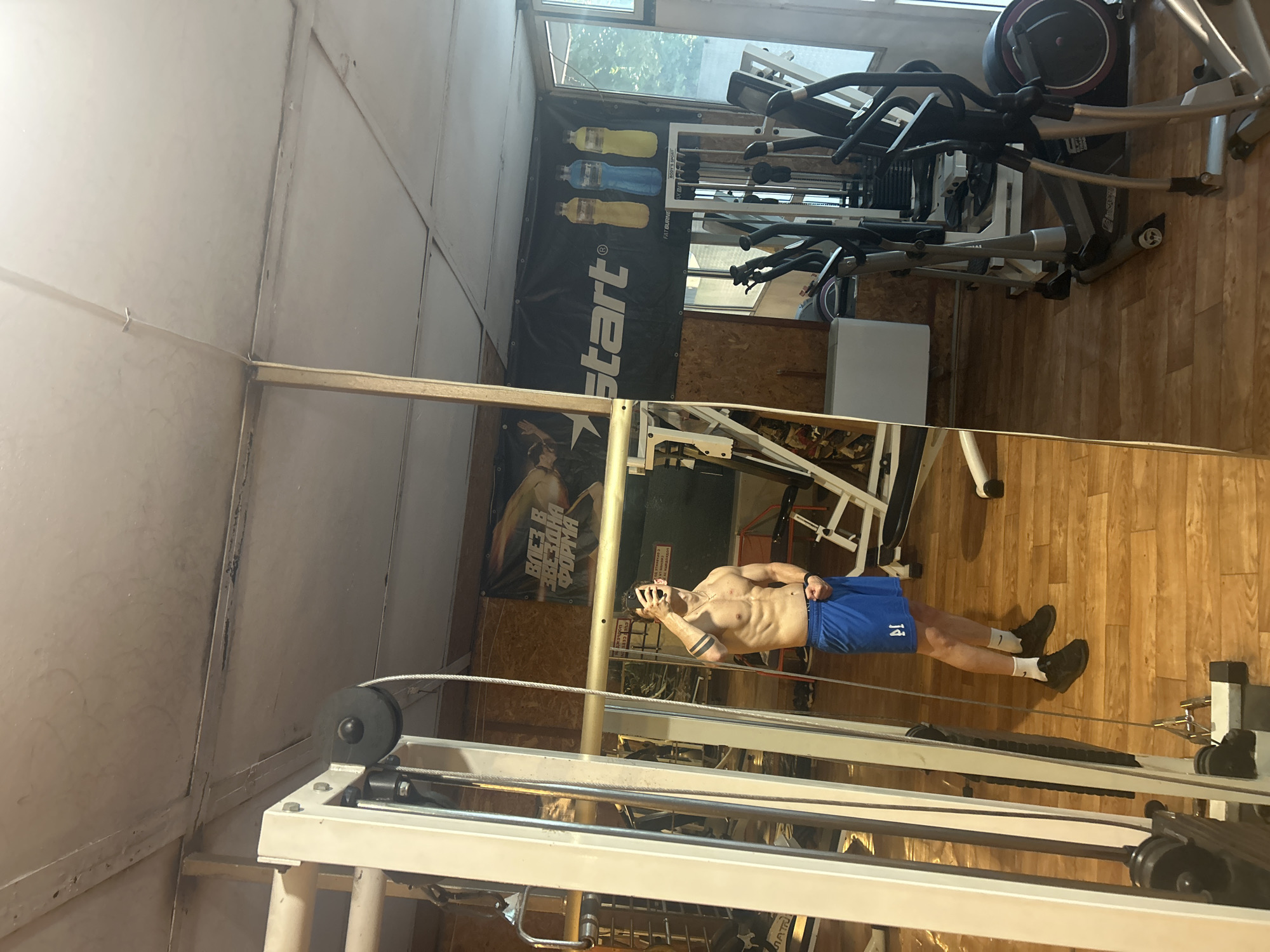 The image depicts the interior of a gym in Elhovo, Bulgaria. A partially nude man in blue shorts is visible in the reflection of a mirror, standing near exercise equipment. In the background, exercise machines are arranged on a wooden floor. A banner with the word "start" is affixed to the wall. The room has a window at the top, providing natural light.