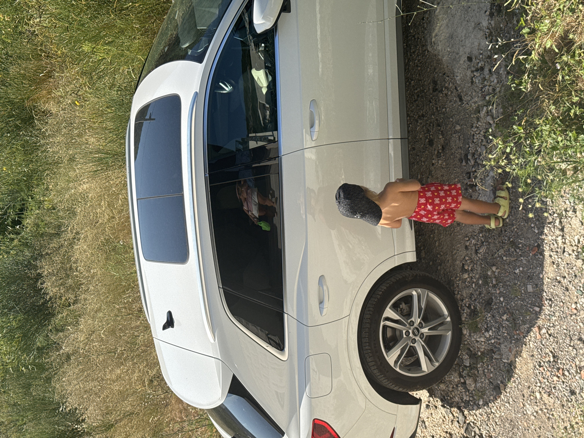 In Rafailovići, Montenegro, a young child wearing a dark patterned hat and red patterned shorts stands on a gravelly ground near a white Audi SUV. The car has a panoramic sunroof and is parked on a dirt road surrounded by dry grasses and green foliage. The child is facing away from the camera, looking towards the vehicle. Reflections are visible in the car windows, showing what appears to be a person inside the vehicle. The scene is bathed in daylight.