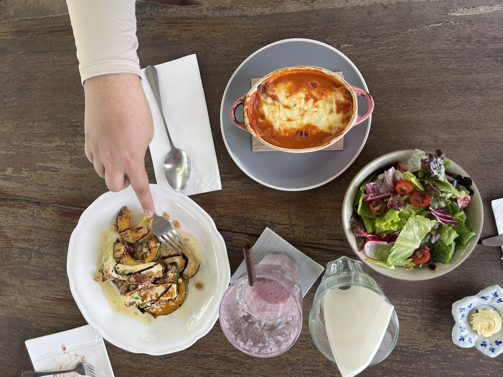 From an overhead perspective, the image depicts a wooden table in what appears to be a restaurant in Ban Tom, Thailand. A person's hand and forearm, clad in a light beige long-sleeved shirt, is visible on the left. The hand holds a fork, poised to spear a piece of food on a white plate. The plate contains a dish of what appears to be roasted squash, halloumi cheese, and other ingredients, drizzled with a sauce or dressing. A small, soiled side plate is also present on the left.
To the right of the squash dish is an empty glass with a straw, remnants of a pink smoothie visible inside. Next to the glass is a jug containing folded white napkins. Adjacent to the jug is a bowl of green salad with tomatoes and radish. At the top right of the image, there's a plate with a gratin dish, with a melted white topping over a tomato-based sauce. A gray plate and a napkin also accompany the gratin dish. A spoon is on the napkin. A small blue and white dish, containing what appears to be butter, is visible on the lower right corner of the table.