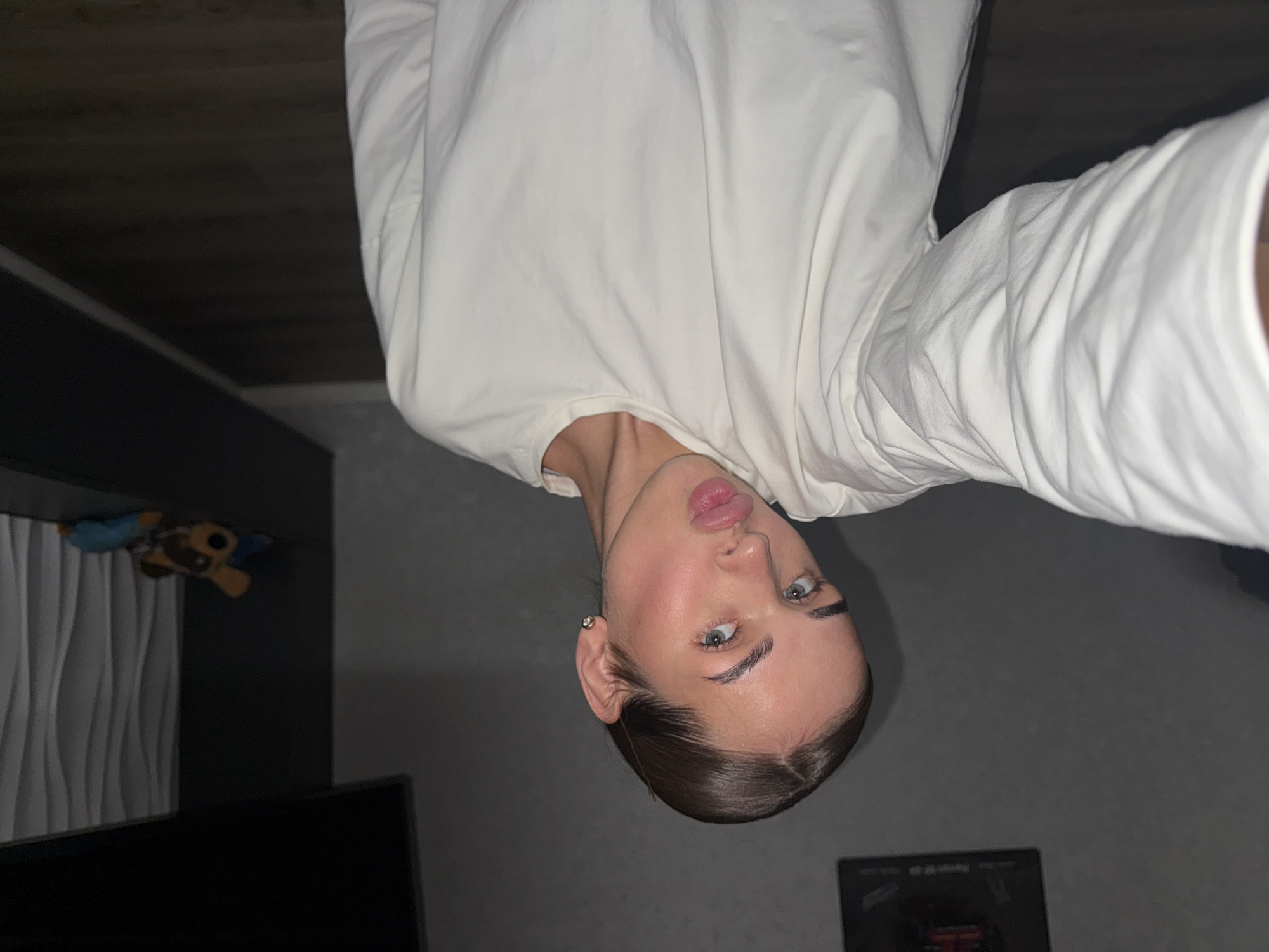 A selfie, taken indoors, showing a fair-skinned individual with light eyes, wearing a white long-sleeved shirt. The background shows a dark grey wall, a shelf with a teddy bear, and an unidentified object on the floor. It is impossible to confirm that the location is in the city of Лобаново, Russia.