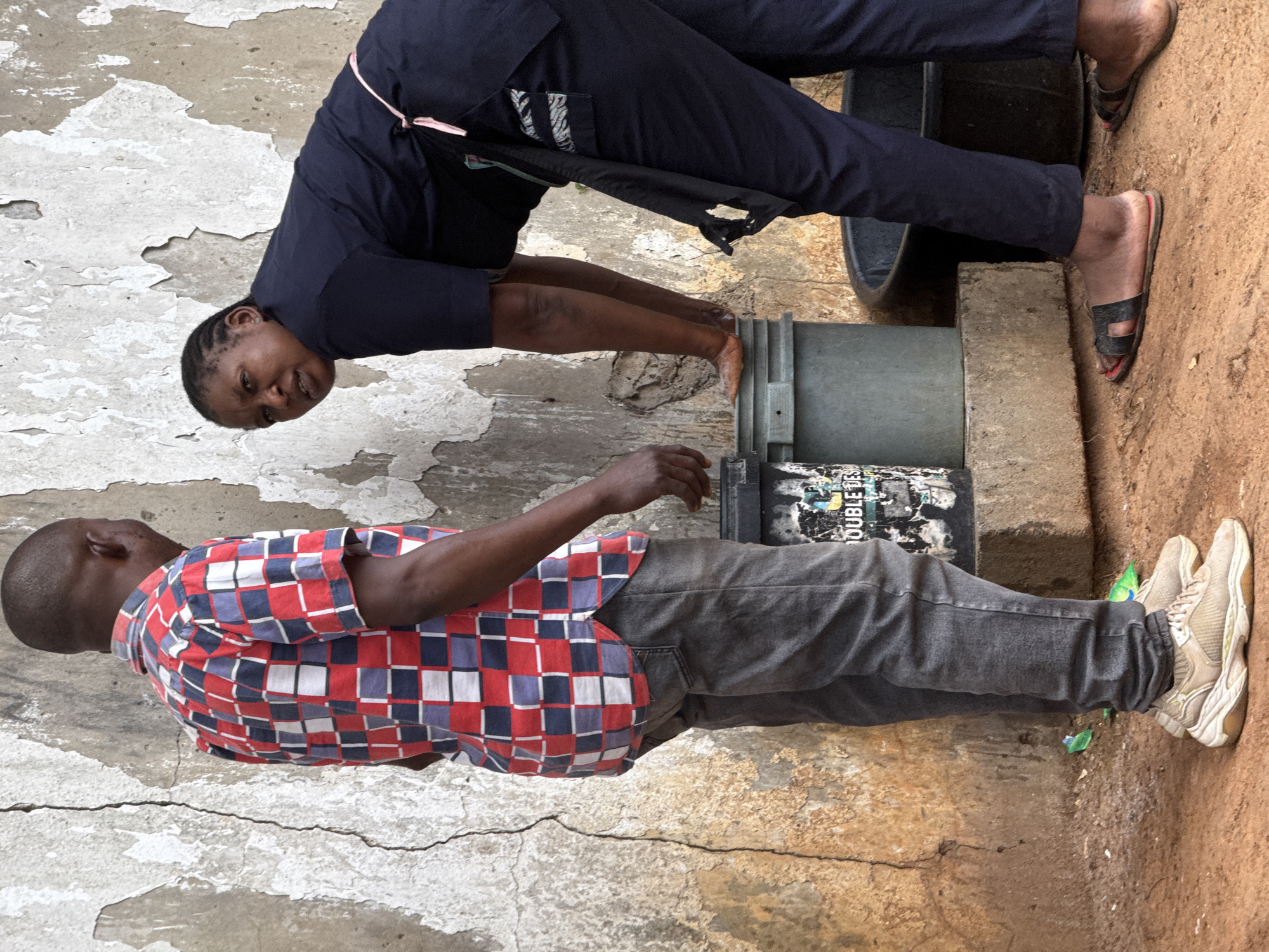 Two people are interacting outdoors in an area that appears to be in a state of disrepair.
One person, a dark-skinned woman, is bending forward and appears to be handling an object of cylindrical shape such as a container. She is wearing a dark blue long-sleeved shirt and dark pants. She has her hair braided. She is wearing open-toed slip-on sandals.
The second person, a dark-skinned man, is standing to the side. He is wearing a short-sleeved shirt with a red, white, and blue checkered pattern, and gray jeans. He has short hair. He is wearing white sneakers.
The environment includes a wall with peeling paint, concrete blocks, a black plastic container, and what looks like dirt ground. The scene is taking place in Abuja, Nigeria.