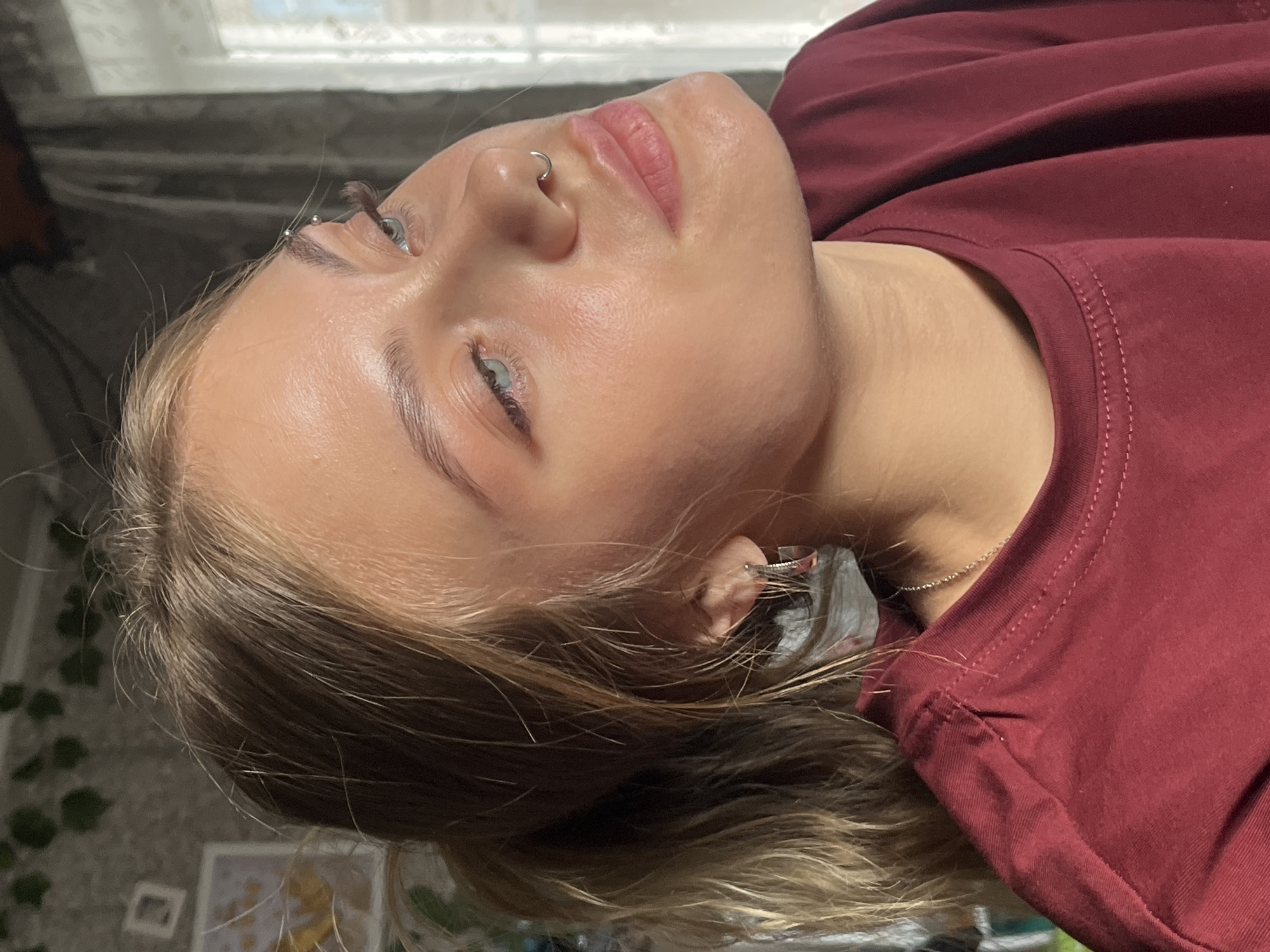 The primary image is a close-up portrait of a young person, presented in a rotated orientation suggesting they are lying down or leaning back. The subject has fair skin, light brown hair with some lighter highlights, and light blue or greenish eyes. They sport a silver hoop septum piercing and a silver stud eyebrow piercing above their left eye (viewer's right). Their lips are naturally pink, and their expression is calm and neutral as they gaze slightly upwards. A thin silver chain necklace is visible around their neck.
The person is wearing a simple maroon or burgundy t-shirt with a visible ribbed collar and stitched seams. Their wavy hair falls around their shoulders, partially obscuring their right ear, where a silver ear piercing may be visible.
The background is softly blurred, indicating a shallow depth of field. Natural light streams from a window in the upper left, illuminating the subject's face. Behind them, a dark, textured fabric, possibly a blanket or curtain, is visible. Further in the background, there are hints of green, ivy-like decorations on a wall and blurred white frames or objects on a surface, suggesting an indoor setting like a bedroom or living space. The soft, natural lighting indicates it is daytime. There is no discernible text within the image. The location context provided is Фоки, Russia.