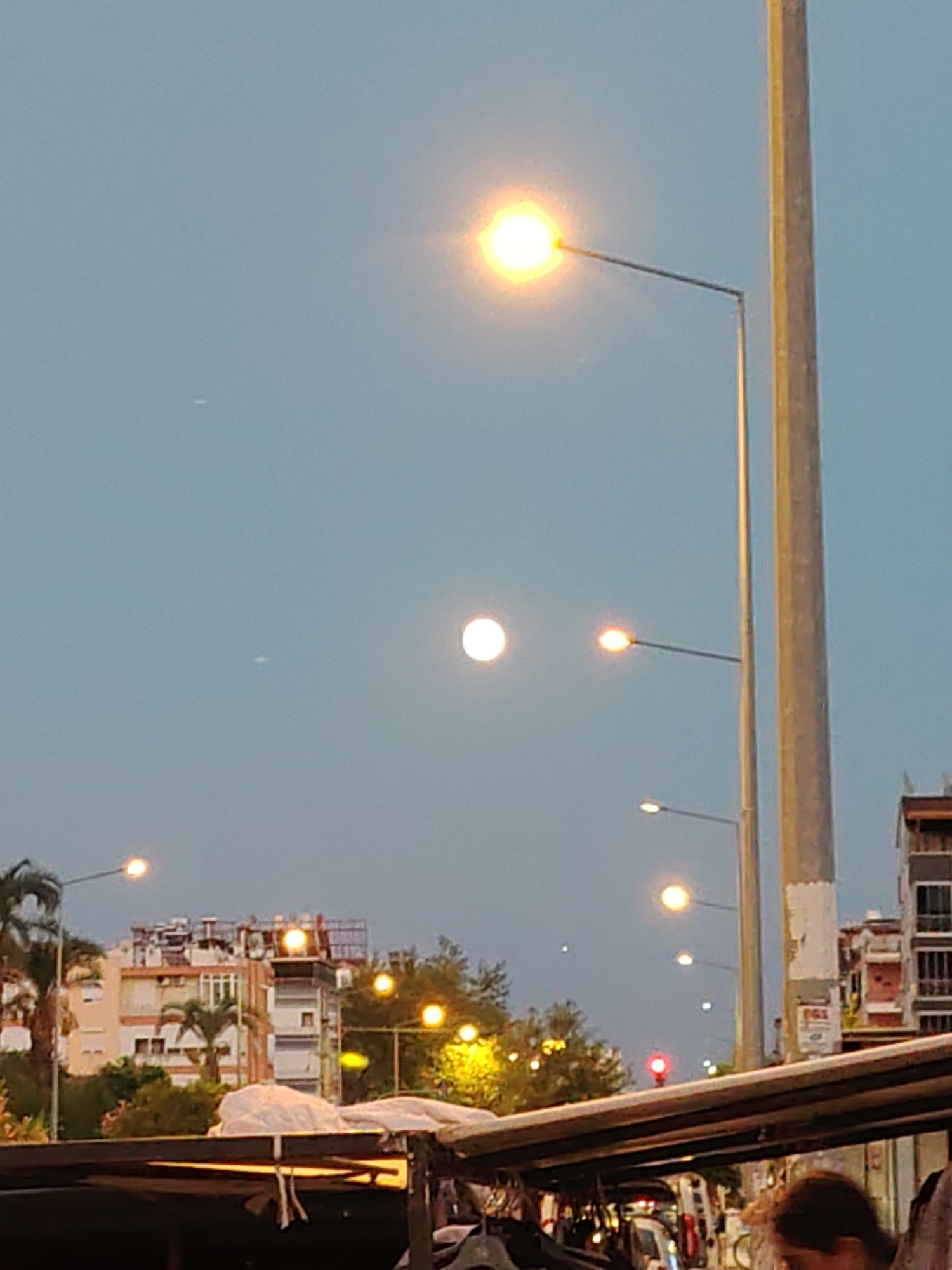 The image depicts a street scene in Konyaaltı, Turkey, during what appears to be twilight. A bright light, possibly the moon or a street lamp, is visible in the upper portion of the sky. Several other streetlights are also illuminated along the road. In the background, buildings with a few palm trees are visible, suggesting a coastal or urban environment. In the foreground, there is a structure that appears to be a market stall or an awning, with some fabric or tarpaulin covering it. Part of a person's head and shoulder is visible in the lower right corner, indicating a human presence. The overall impression is that of a public space transitioning from day to night.