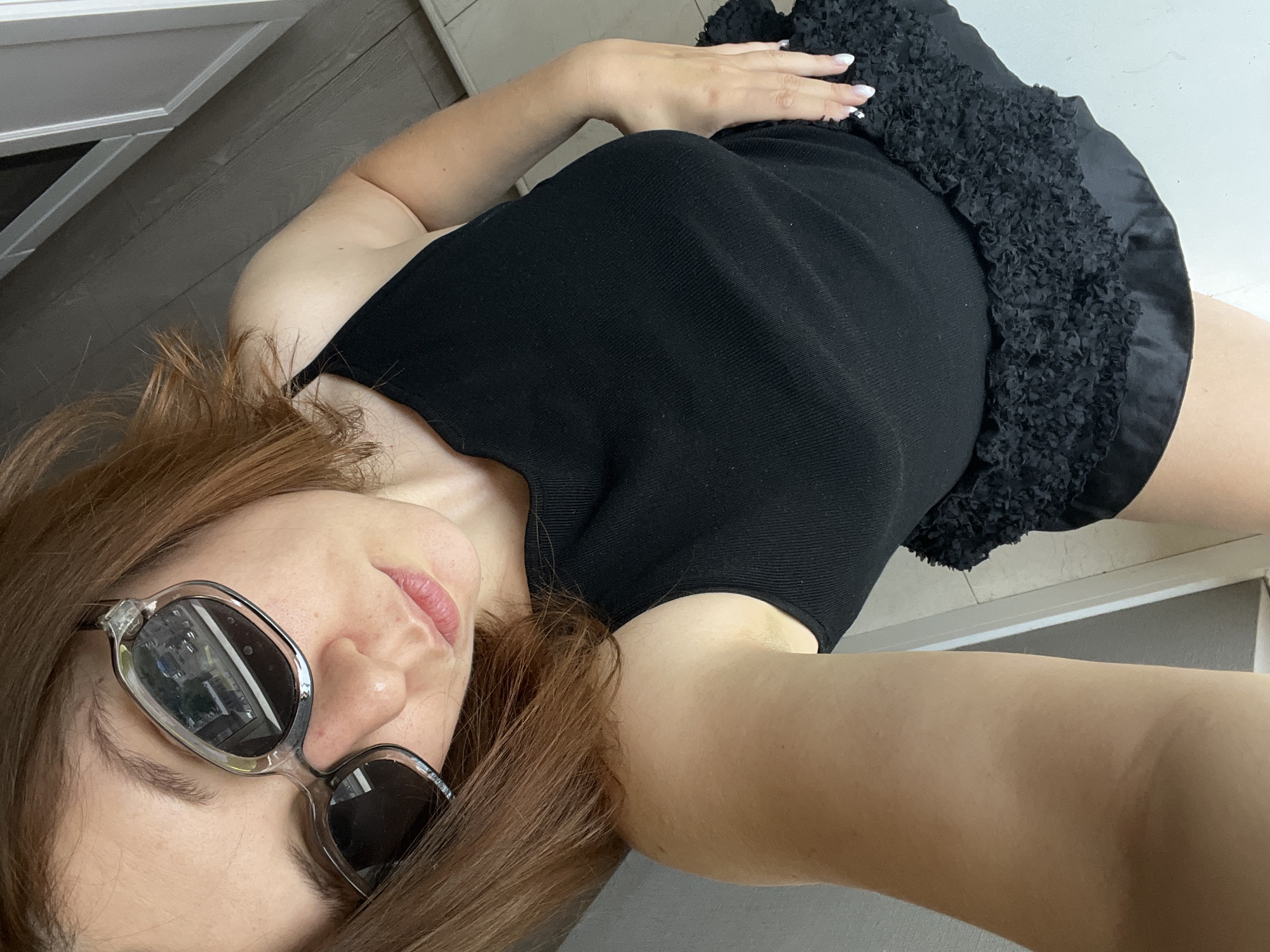 A woman with auburn hair and fair skin is taking a selfie. She is wearing a black tank top and a black ruffled skirt. She is also wearing sunglasses with clear frames. The background appears to be an outdoor setting, possibly a balcony or patio, with wooden flooring and a light-colored wall or railing. The image quality is decent, with good lighting. There is no information in the image that indicates the location is the city of Krasnoyarsk, Russia.