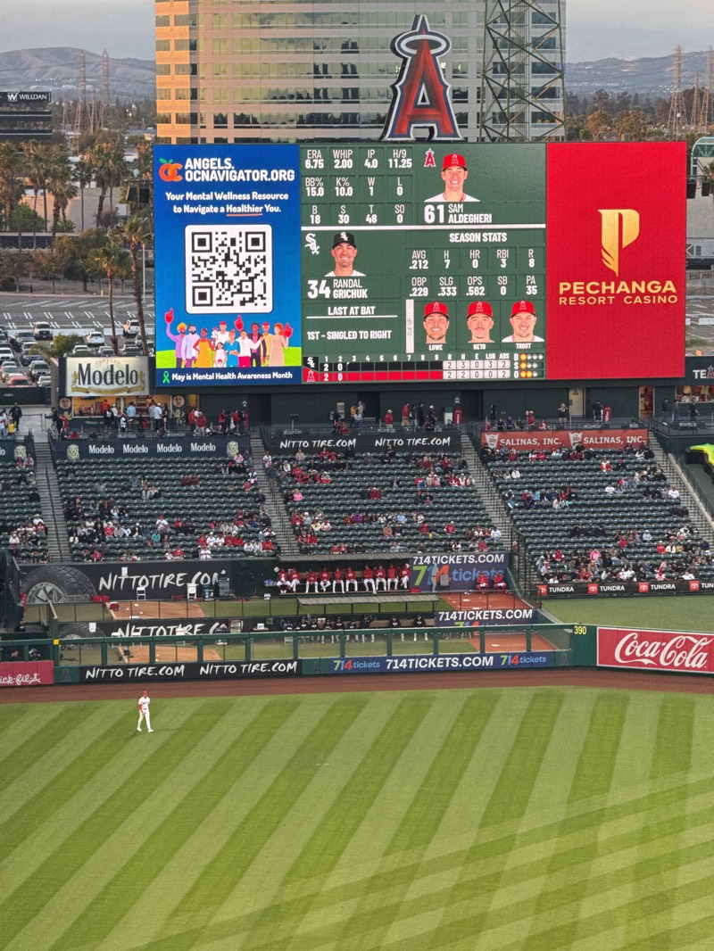 The image captures a baseball game in progress at Angel Stadium in Anaheim, California, United States. The focus is on the stadium's large video screen, displaying team statistics and advertisements. On the left side of the screen, a blue advertisement with the text "ANGELS. OCNAVIGATOR.ORG" promotes mental wellness resources and includes a QR code. To the right, baseball statistics for players like Randal Grichuk and Sam Alldegheri are shown. A red advertisement for Pechanga Resort Casino occupies the right side of the screen.
Below the video screen, spectators are seated in the stands, watching the game. Advertisements from companies such as Modelo, Nitto Tire, and 714Tickets line the stadium walls. On the baseball field, a single player in a white uniform stands on the field. The green grass is precisely striped. Palm trees and buildings can be seen in the background, suggesting the stadium's urban setting.