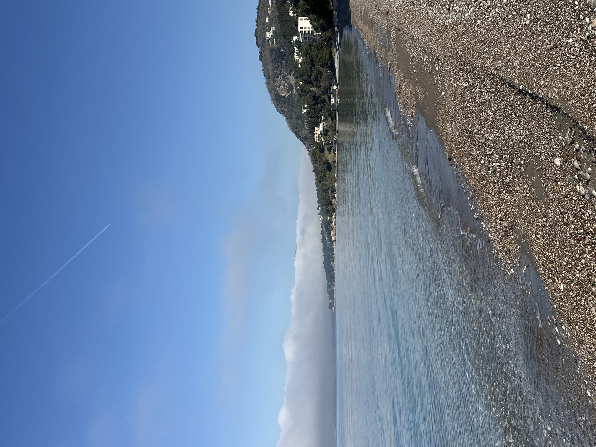 The image depicts a coastal scene in Šušanj, Montenegro. A pebble beach is seen on the right, transitioning to calm, clear water. Along the shore, there's a tree covered hill. There are houses and/or buildings built on the hillside. A thin layer of fog or low-lying clouds obscures the horizon between the water and the sky. Above the fog, a clear, blue sky is visible, with a contrail of an airplane.