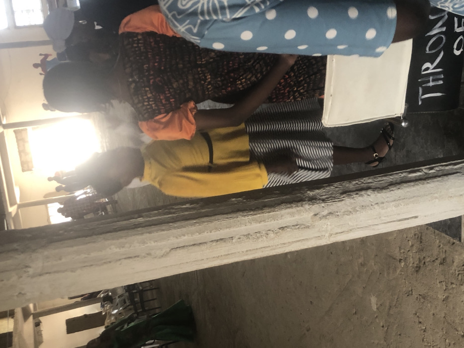 The image is rotated 90 degrees clockwise. When viewed upright, it shows an indoor scene with several people, focusing on two individuals in the mid-ground.
In the center of the frame, a person is visible from the chest down, wearing a yellow short-sleeved top and a vertically striped skirt (dark and light stripes, possibly black and white or grey and white). Their right arm is bent at the elbow, with their hand out of frame to the left. They are wearing black open-toed sandals with ankle straps on their feet. To their immediate right, at about knee height, there is a white, rectangular object, possibly a small table, chair, or bag.
Slightly behind and to the left of this person, another individual is seen, wearing an orange short-sleeved top layered under a dark, possibly patterned or textured, long-sleeved dress or tunic. Their head and upper torso are visible, with dark hair.
In the upper right corner of the image, the leg or lower body of a third person is partially visible, clad in light blue fabric with white polka dots.
In the background, multiple indistinct figures are visible, silhouetted against a very bright, overexposed light source, which appears to be a window or a set of windows. This bright light makes it difficult to discern details of the background figures or the architecture of the far wall. The overall setting appears to be a large indoor space, possibly a hall, with visible concrete beams or ledges in the foreground and lower mid-ground.
To the right of the person in the yellow shirt and behind the white object, a black sign stands upright. White text is visible on the sign, which appears to read "THRONES OF".
The activity seems to be an indoor gathering or event, with people standing and possibly observing. The lighting suggests either bright daylight coming from windows or strong artificial indoor lighting. The overall atmosphere appears casual.
