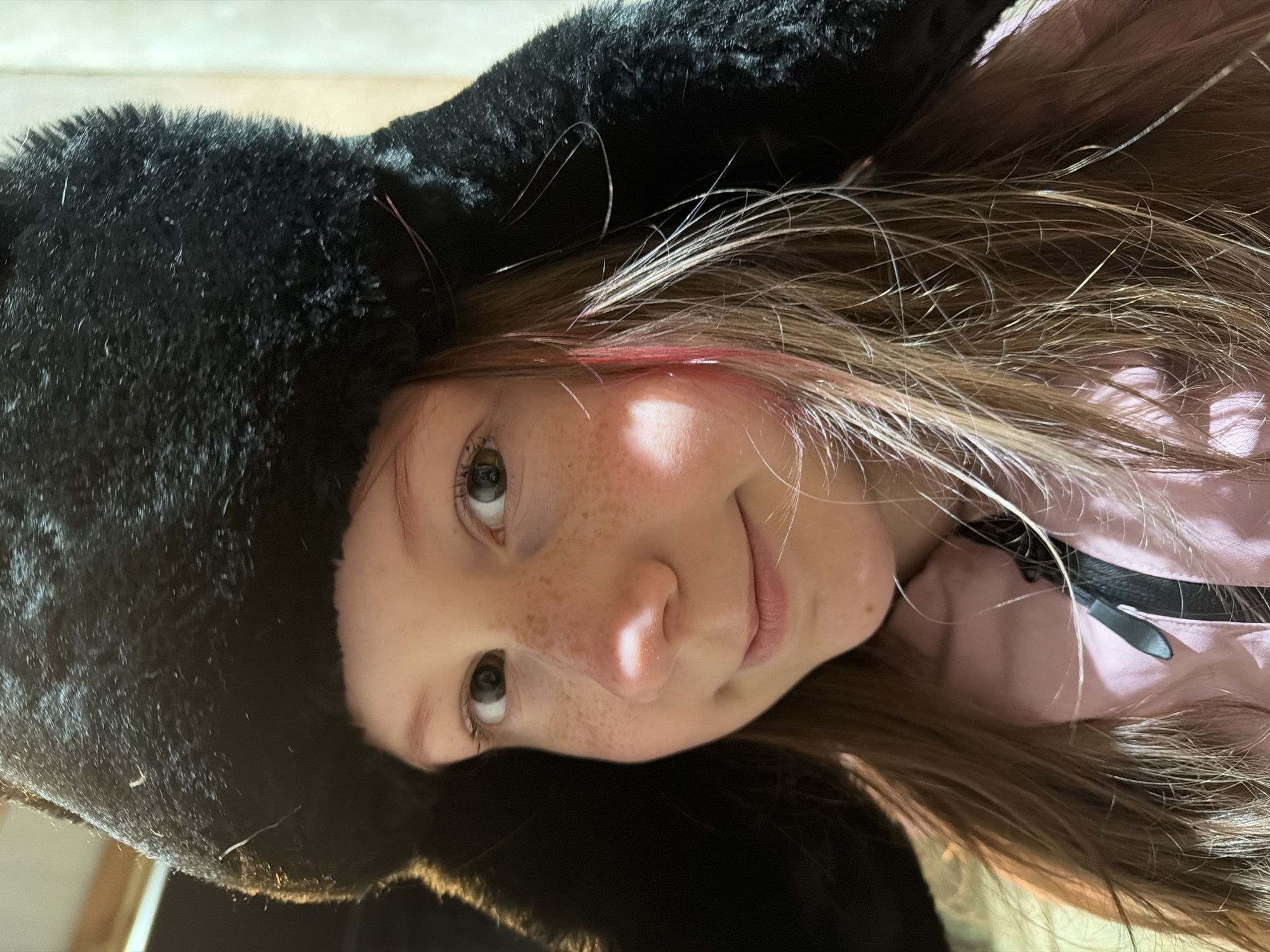 A close-up portrait depicts a young individual, likely a child or pre-teen, looking directly at the camera with a slight smirk. The image is rotated 90 degrees counter-clockwise, showing the person's head tilted to the right. They have light brown hair, notable for several bright pink or red strands, and prominent freckles scattered across their nose and cheeks. Their eyes are light in color. The person is wearing a large, fluffy black hat, possibly made of faux fur, which frames their face. Below the hat, a pink garment, possibly a jacket or top, is visible, featuring a black zipper. The lighting illuminates the person's face from the left side (as oriented in the image), suggesting an indoor setting. There is no visible text in the image. While the context provided indicates Markovo, Russia, there are no specific visual cues within the image to confirm this location.