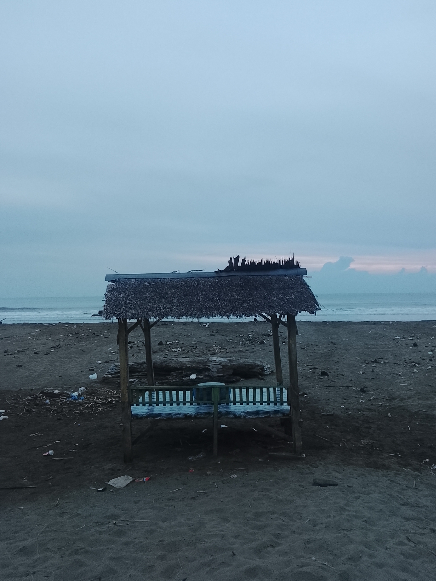 A simple wooden swing set with a thatched roof stands on a dark sand beach, facing the ocean under an overcast sky. The beach is littered with debris, including plastic waste and driftwood. In the background, waves gently roll onto the shore, and the horizon is marked by a muted, dusky sky with faint hints of pink near the water. The overall impression is one of a somewhat neglected and desolate coastal scene, likely in Reudeuep, Indonesia, given its typical appearance. There are no people visible in the image.