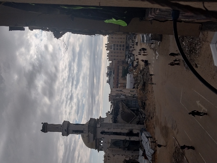 The image depicts a devastated urban landscape with a damaged mosque minaret prominently displayed. Buildings are reduced to rubble, and debris litters the streets. Several people can be seen moving through the area, perhaps assessing the damage or seeking safety. The sky is overcast, adding to the somber and desolate atmosphere. The perspective suggests the photo was taken from a high vantage point, overlooking the destruction.
