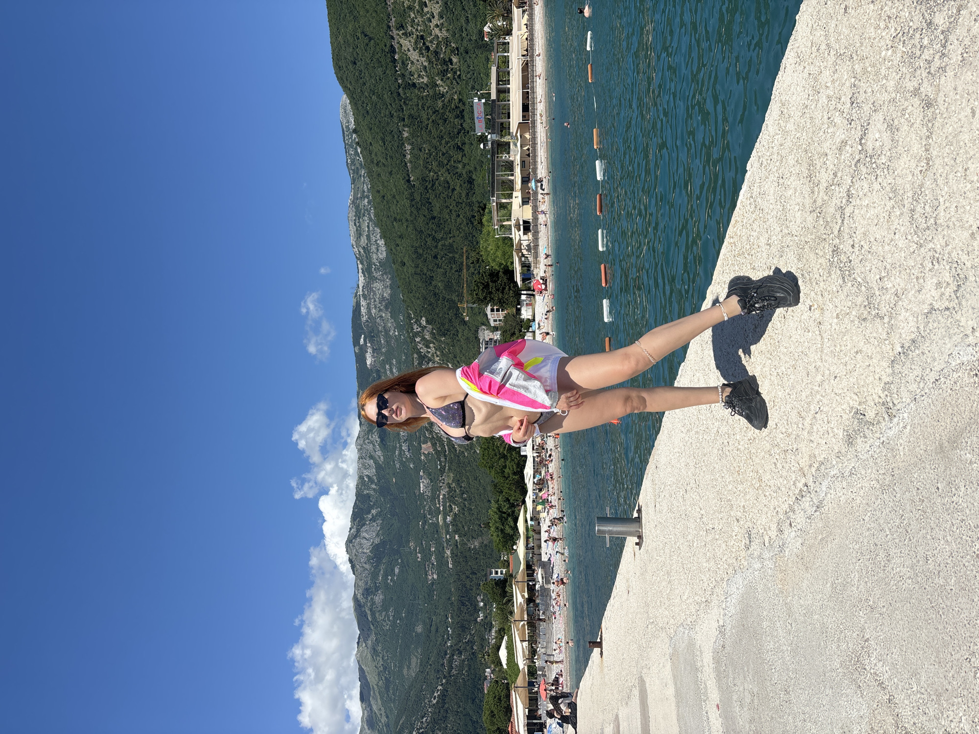 A young woman with reddish-brown hair and sunglasses stands posed on a textured concrete pier or wall, looking towards the camera. She is wearing a sparkly bikini top, white shorts, a bright pink, white, and silver jacket, and black athletic shoes, with delicate bracelets visible on her wrists.
The pier extends into calm, clear turquoise water, marked by a line of orange and white buoys. In the mid-ground, a busy beach stretches along the coastline of Budva, Montenegro, populated by many people sunbathing and swimming. The beach features numerous sun loungers, umbrellas, and several multi-story buildings, one of which prominently displays the word "BOCCA."
Behind the beach, lush green, forested mountains rise dramatically, dominating the background under a vibrant blue sky with a few small, scattered white clouds. The bright sunshine and clear sky suggest it is midday or early afternoon. The scene captures a lively coastal holiday atmosphere, with the woman enjoying the scenic resort setting.