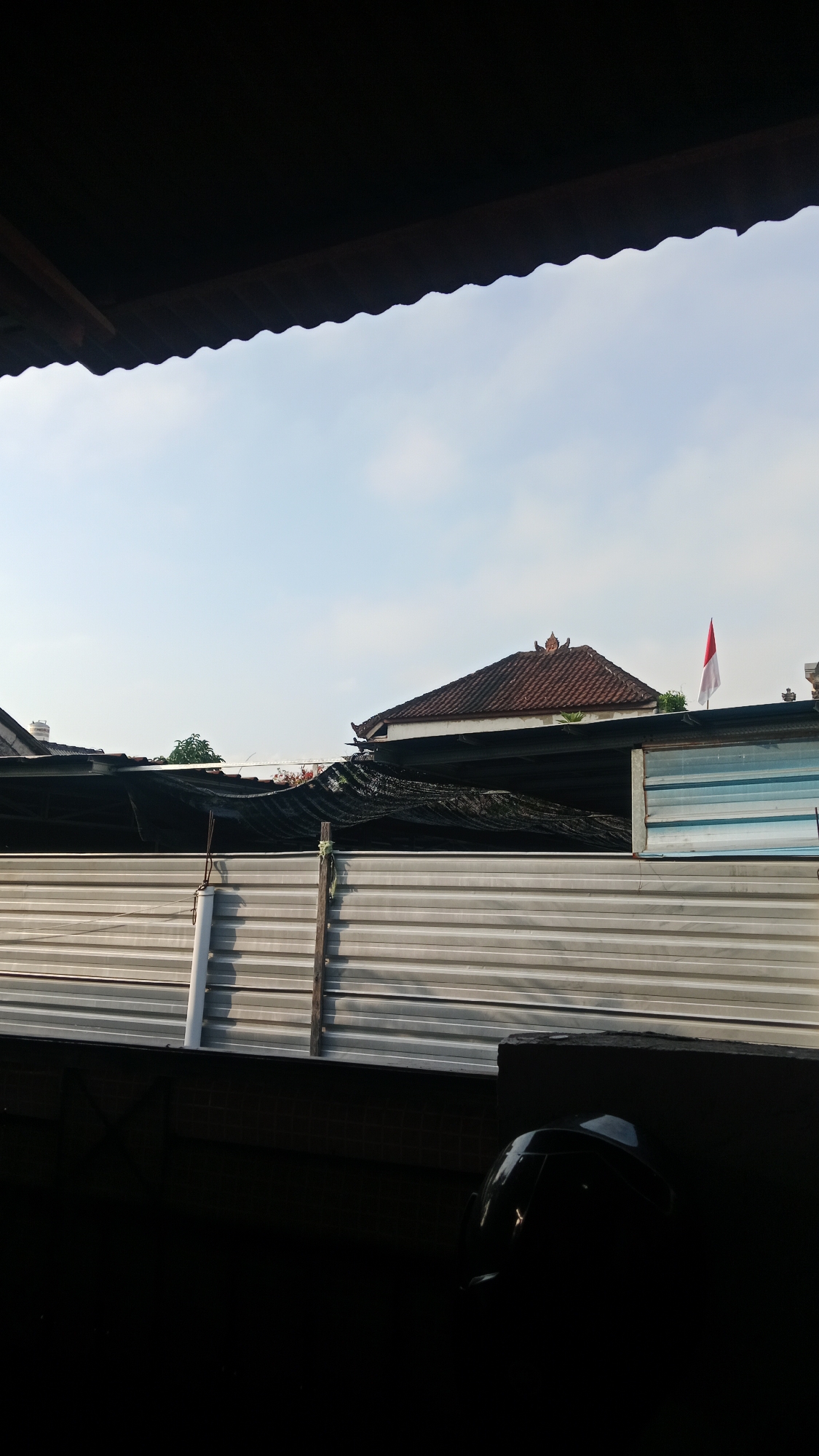 The image captures an outdoor scene under a clear sky, possibly in the city of Denpasar, Indonesia. In the foreground, a dark, silhouetted area suggests the presence of a shaded overhang or structure, with the edge of corrugated roofing visible. Below this, a motorcycle helmet is partially visible in the darkness.
Beyond the foreground, a corrugated metal fence stretches across the midground, separating different areas. Behind the fence, several structures are visible. One is a house with a traditional tiled roof, featuring intricate architectural details at its apex. A red and white flag, likely the Indonesian flag, is flying from a pole atop a nearby structure. Another building with a dark, possibly tarpaulin-covered roof, is positioned in front of the house. The overall impression is of a residential or semi-industrial area, with a mix of building materials and styles. The lighting suggests it is daytime, possibly morning or late afternoon, given the angle of the shadows.