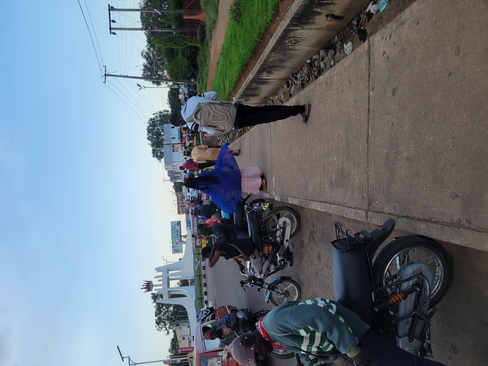 A street scene in Barakin Lerawi, Nigeria, captures a busy intersection with various modes of transportation and pedestrians. In the foreground, a man wearing a green and white striped hoodie and a grey beanie sits on a motorcycle. To his right, another motorcycle is occupied by a man with dreadlocks wearing a black t-shirt and grey shorts. Behind them, a woman in a vibrant blue dress and a pink skirt walks away from the camera, followed by a man in a light blue shirt and black pants carrying a beige backpack.
The background reveals a prominent white monument with a red crown on top, likely a landmark in the city. Several other motorcycles and a red vehicle are visible, suggesting a bustling commercial or transportation hub. Power lines crisscross the sky, supported by wooden poles. The sidewalk area is lined with a concrete barrier bordering a ditch filled with litter. Lush green grass grows on the other side of the ditch, and trees are scattered throughout the environment. The sky is a clear, pale blue, indicating daylight conditions. The scene depicts a typical day in a Nigerian city, characterized by active street life and local culture.