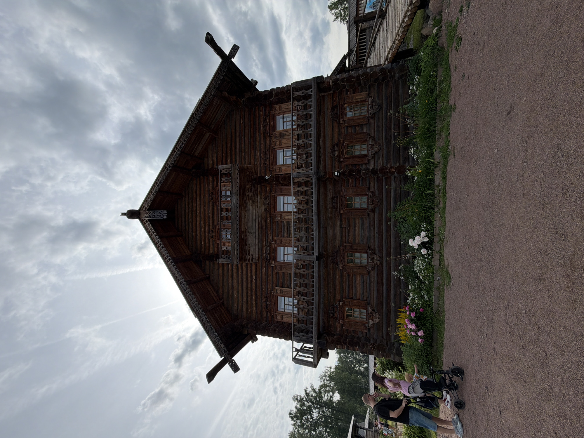 In Dnp Ryzhiki, Russia, a large, ornate wooden house with a steep roof and intricate carvings stands under a cloudy sky. The house has two balconies and multiple windows with wooden shutters. In the foreground, a man in a black t-shirt and denim shorts stands with his arm around a woman in a pink shirt. A child is in a stroller next to them, with the child reaching out towards a flowerbed. The flowerbed contains pink and yellow flowers and greenery. The ground in front of the house is covered in gravel. A wooden ramp leads up to the right side of the house.
